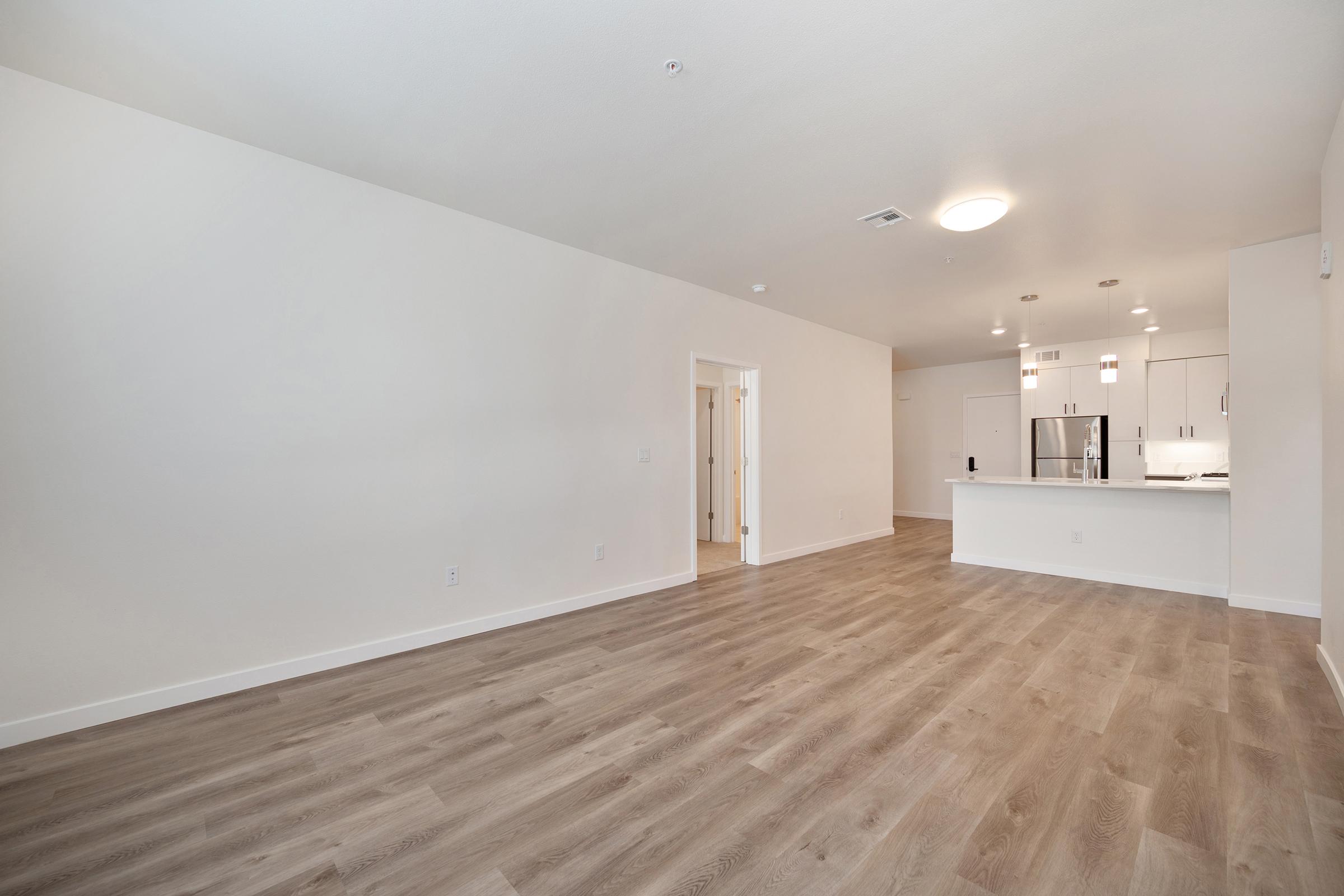 A spacious and modern interior of an empty living area featuring light wood flooring, neutral walls, and a bright atmosphere. The space includes a kitchen with white cabinetry and stainless steel appliances visible in the background, along with doorways leading to other rooms.