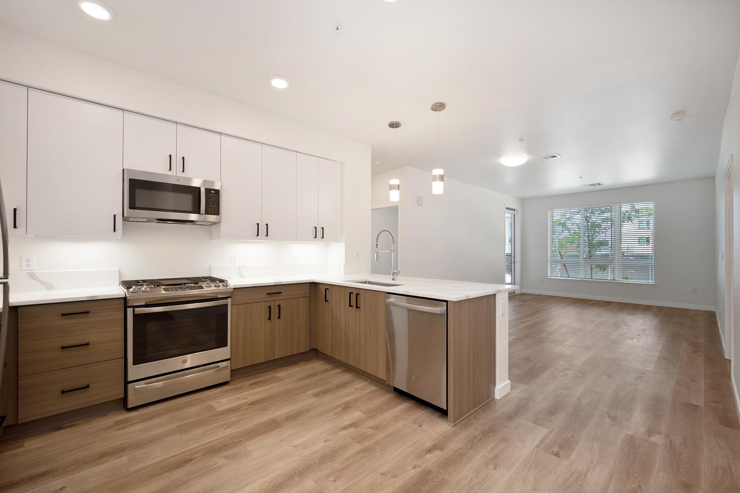 Modern kitchen featuring sleek white cabinets, stainless steel appliances, and a spacious countertop. The open layout leads into a bright living area with large windows showcasing natural light. Hardwood flooring flows throughout, enhancing the contemporary design.