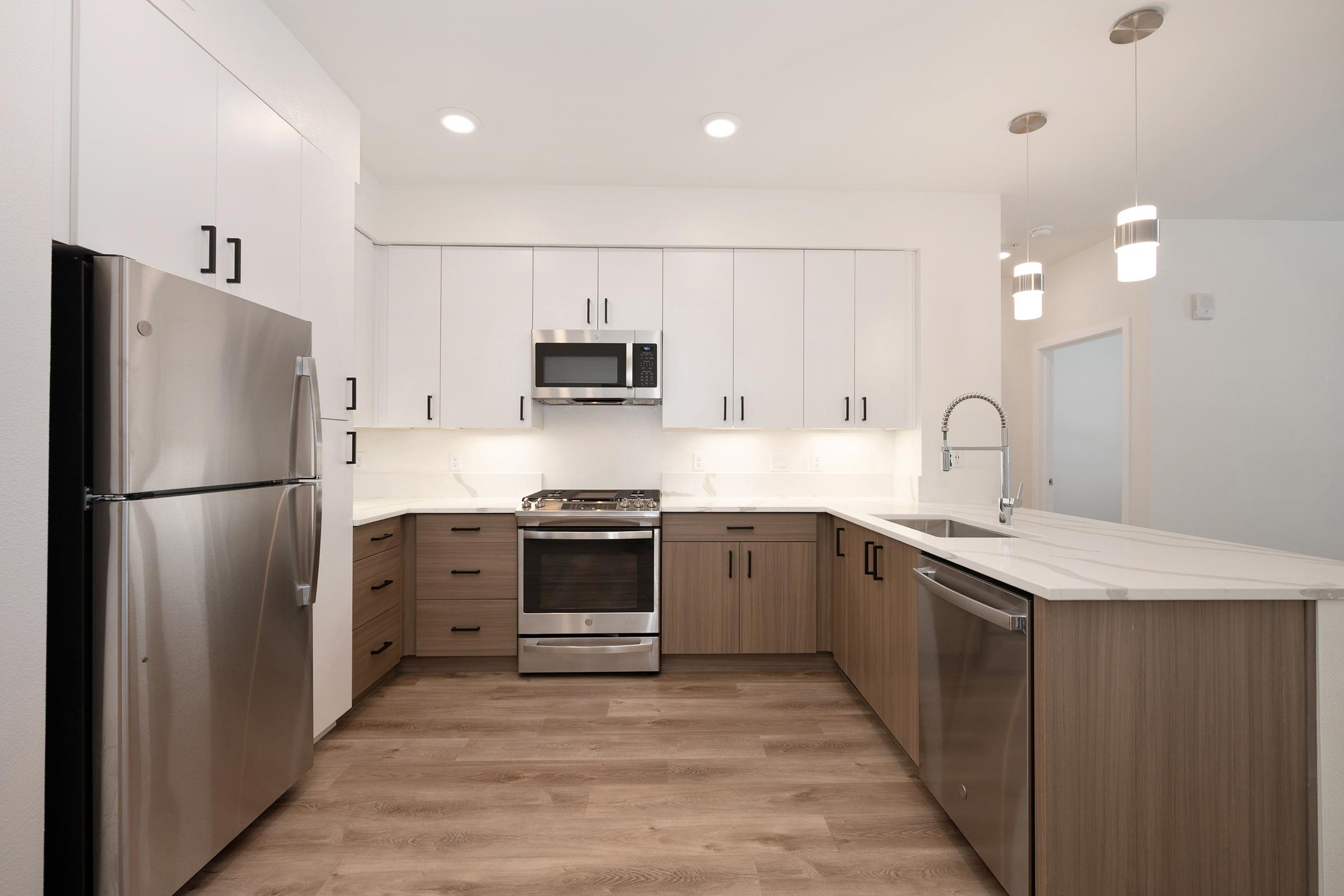Modern kitchen with a sleek design featuring stainless steel appliances, including a refrigerator, microwave, and stove. The cabinetry is a mix of white and wood tones, complemented by a light-colored countertop. Pendant lighting hangs above the island, and the flooring is a warm wood finish.