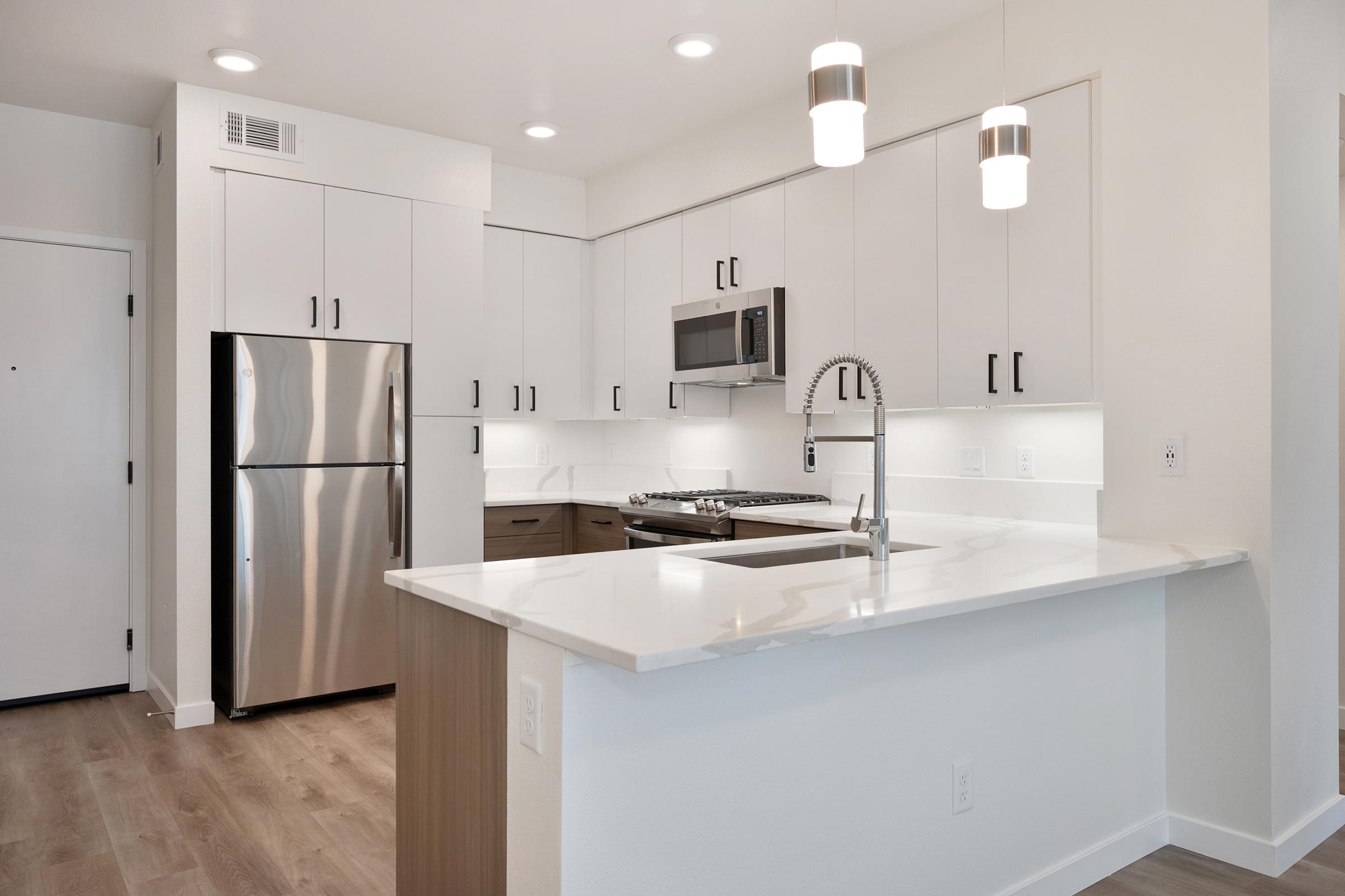Modern kitchen featuring stainless steel appliances, including a refrigerator and microwave. The space has white cabinetry with sleek black handles, a light-colored countertop, and a stylish faucet. Pendant lights hang above the kitchen island, illuminating the open layout with warm wood flooring.