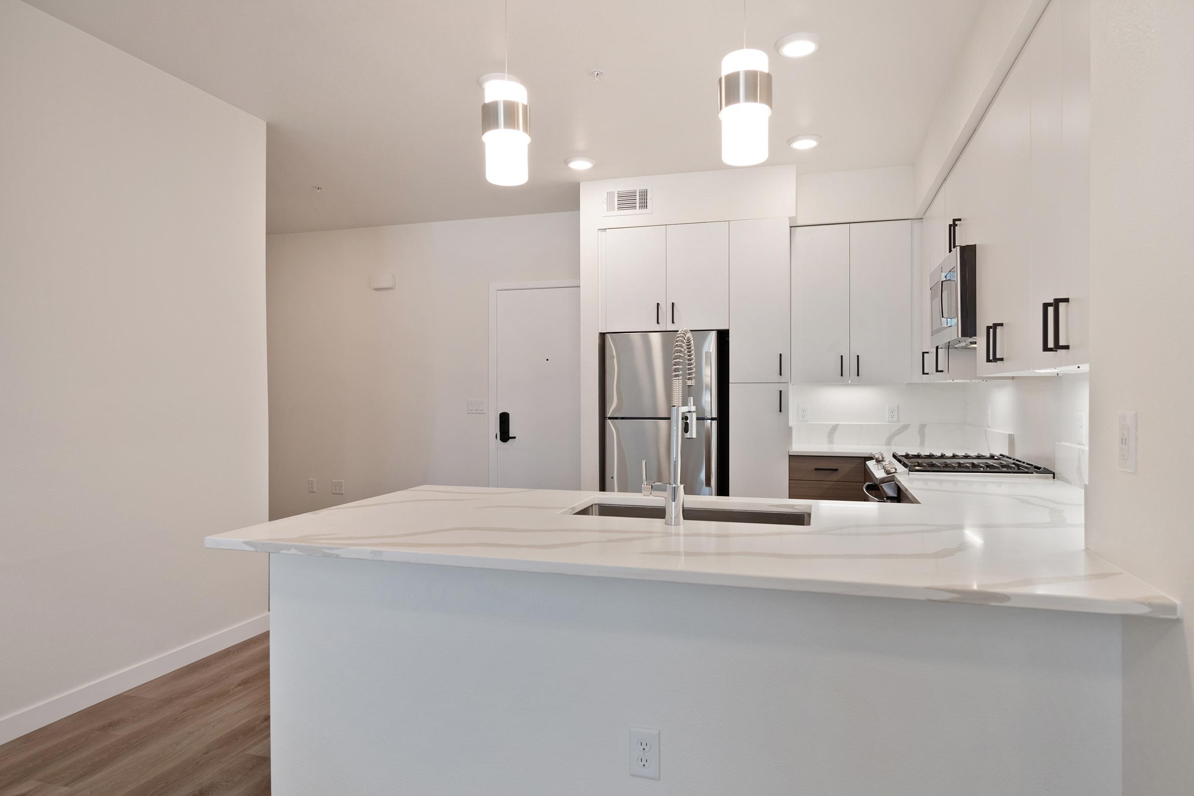 Modern kitchen with white cabinetry, stainless steel appliances, and a large island featuring a sink. The space has pendant lighting and a neutral color palette, creating a clean and bright atmosphere. A doorway on the left leads to an entrance, while the flooring is a light wood finish.