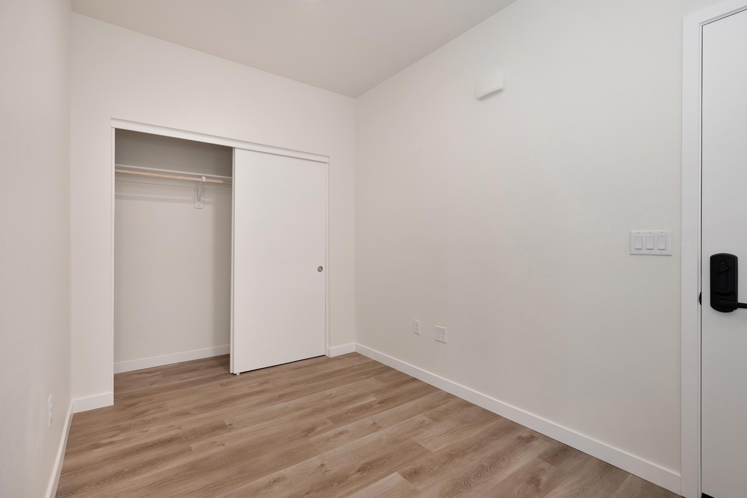 A blank room with light-colored walls and wood-like flooring. There is a sliding closet door on the left, revealing an empty closet. The door to the room features a handle and is on the right side of the image. The space is well-lit and minimalistic, creating a clean and modern aesthetic.