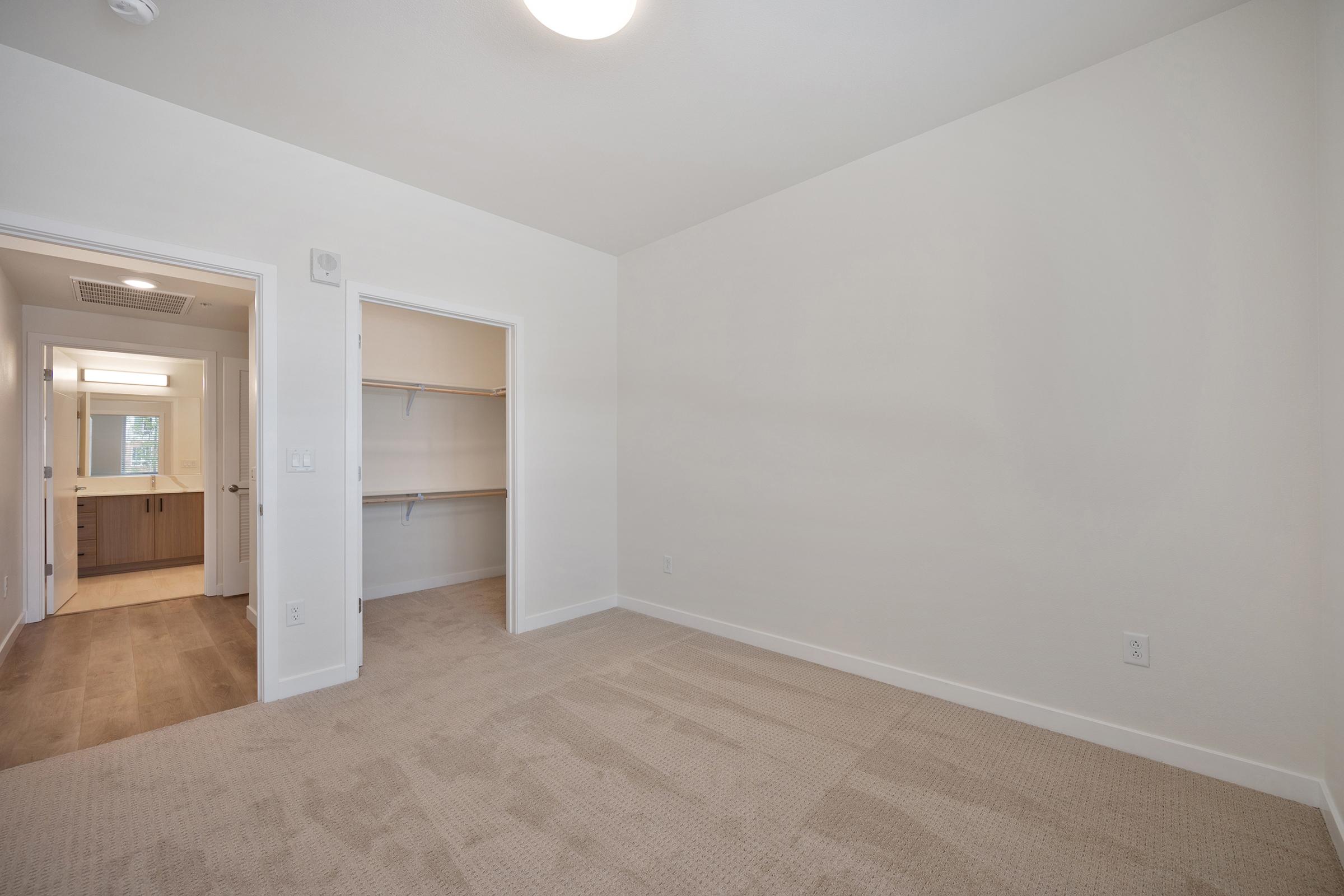 A spacious, empty room with beige carpet and light-colored walls. A doorway leads to a bathroom area, while another door opens to a closet. Natural light fills the room, creating a bright atmosphere with minimal furnishings.