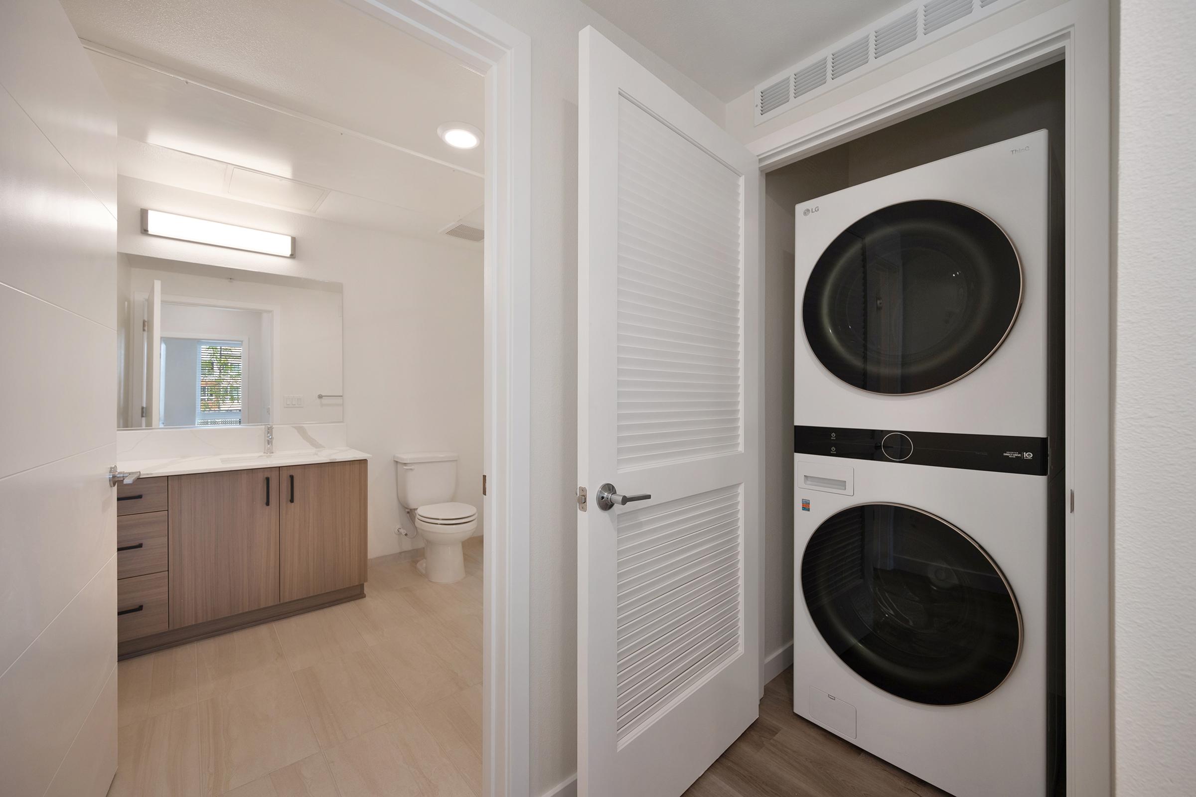 A modern bathroom with a vanity and mirror, featuring a toilet and natural light. Adjacent to the bathroom, there is a laundry area with a stacked washer and dryer, housed in a closet with sliding doors. The interior has light-colored walls and tiled flooring, creating a clean and spacious atmosphere.