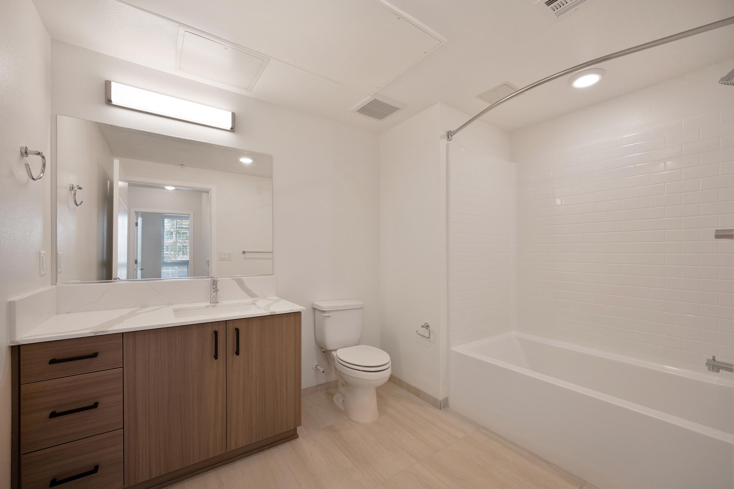 Modern bathroom featuring a sleek design with a large mirror above a double sink vanity, wooden cabinetry, a toilet, and a bathtub with a glass shower enclosure. The walls are tiled in white, and the floor has a neutral light color. Natural light enters through a window in the background.