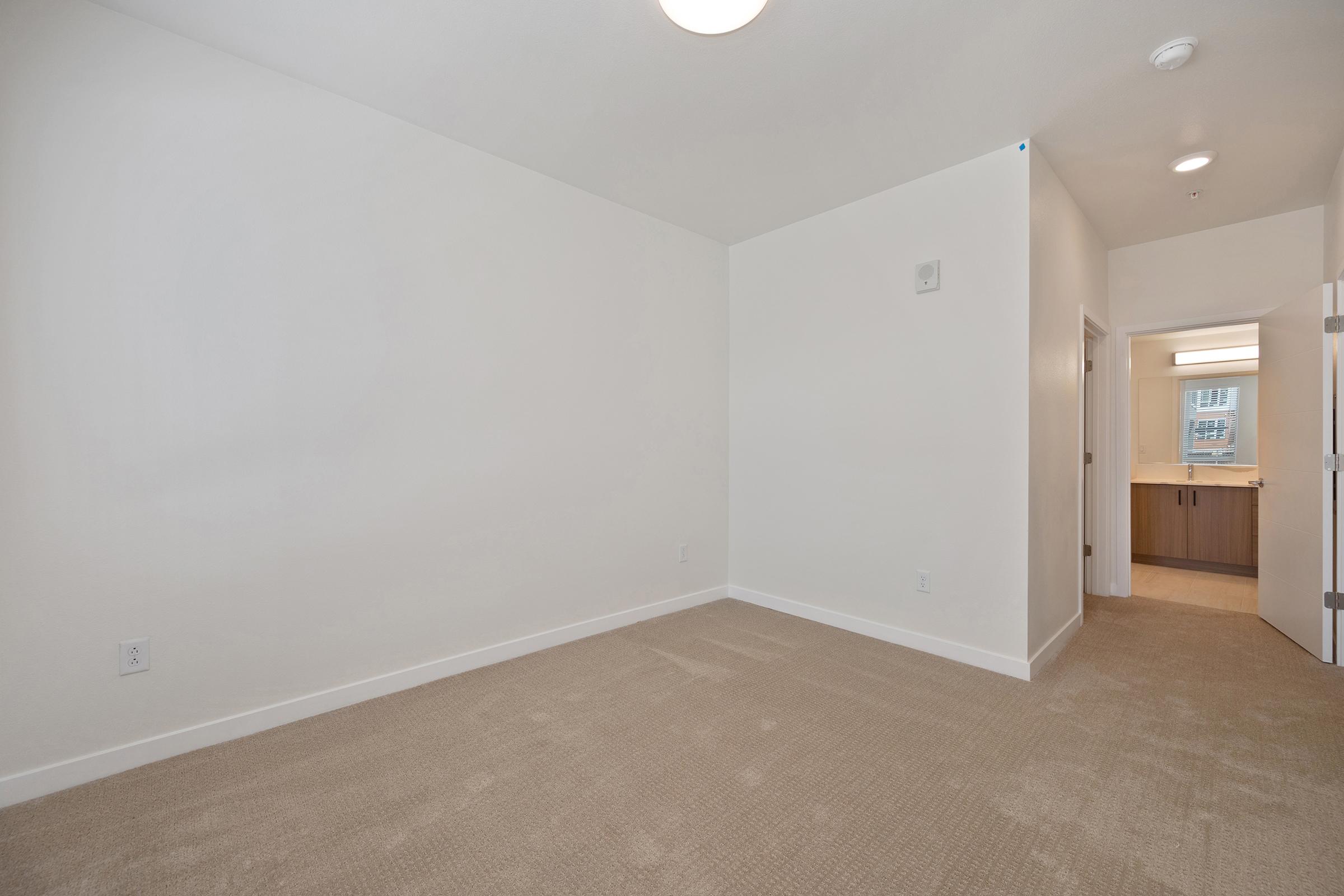 A spacious, empty room with light beige carpet and white walls, featuring a ceiling light. A doorway on the right leads to a bathroom, visible in the background, with modern cabinetry. The room has natural light coming from a window, creating a bright and open atmosphere.