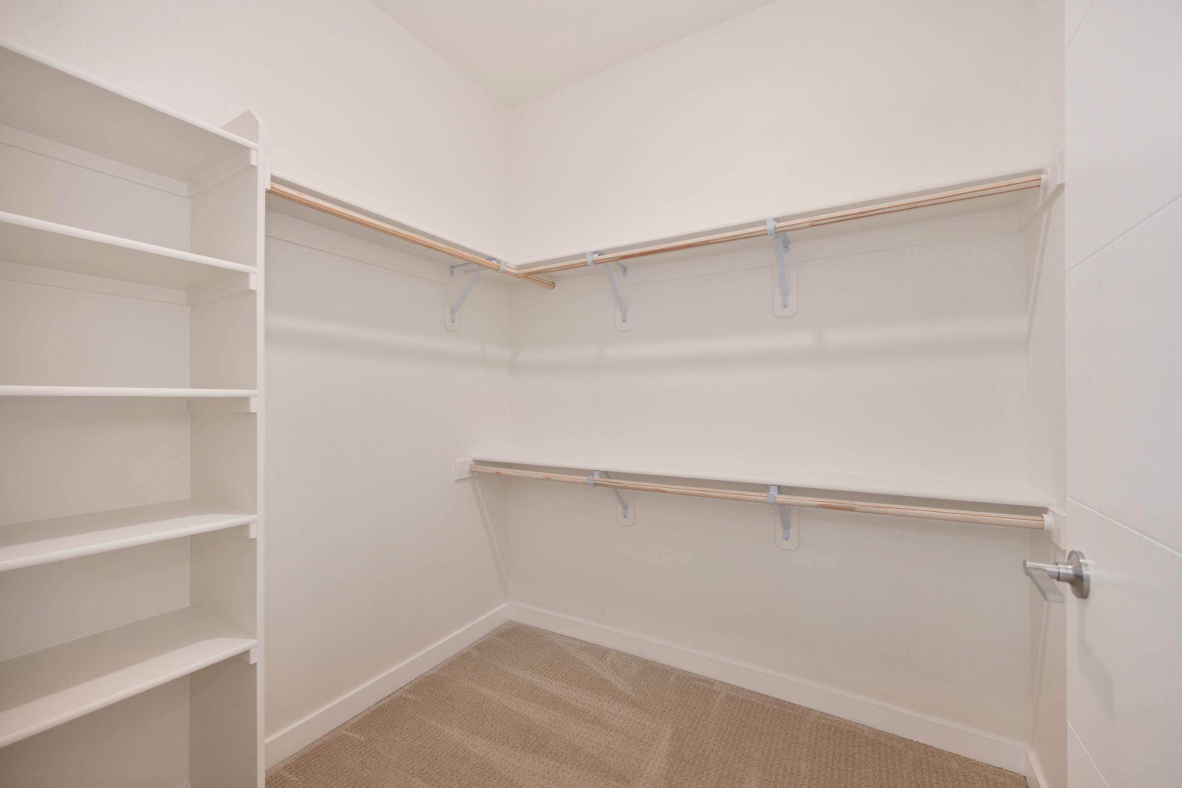 A spacious, empty closet featuring white shelving on the left and two hanging rods along the back wall. The floor is carpeted, and the walls are painted white, creating a clean and modern look. The shelves are unoccupied, highlighting the available storage space.