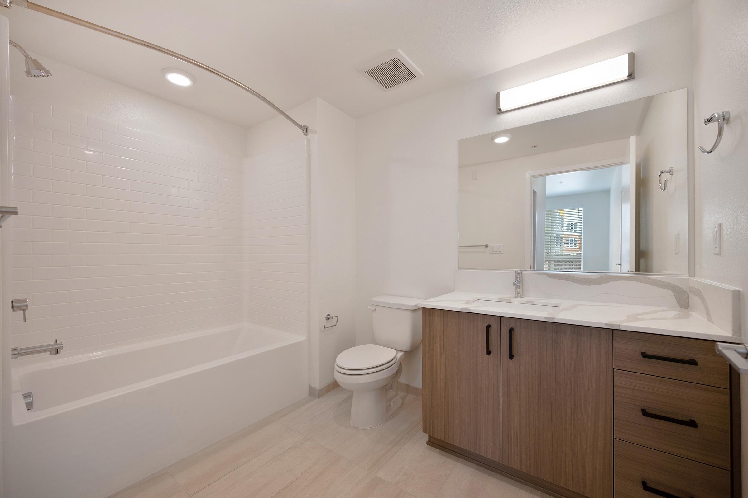 Modern bathroom featuring a white tiled shower and bathtub, a sleek vanity with dark wood cabinetry, a white toilet, and a well-lit mirror. The space is well-organized and has a clean, minimalist design with neutral tones. Soft lighting adds to the overall bright and inviting atmosphere.