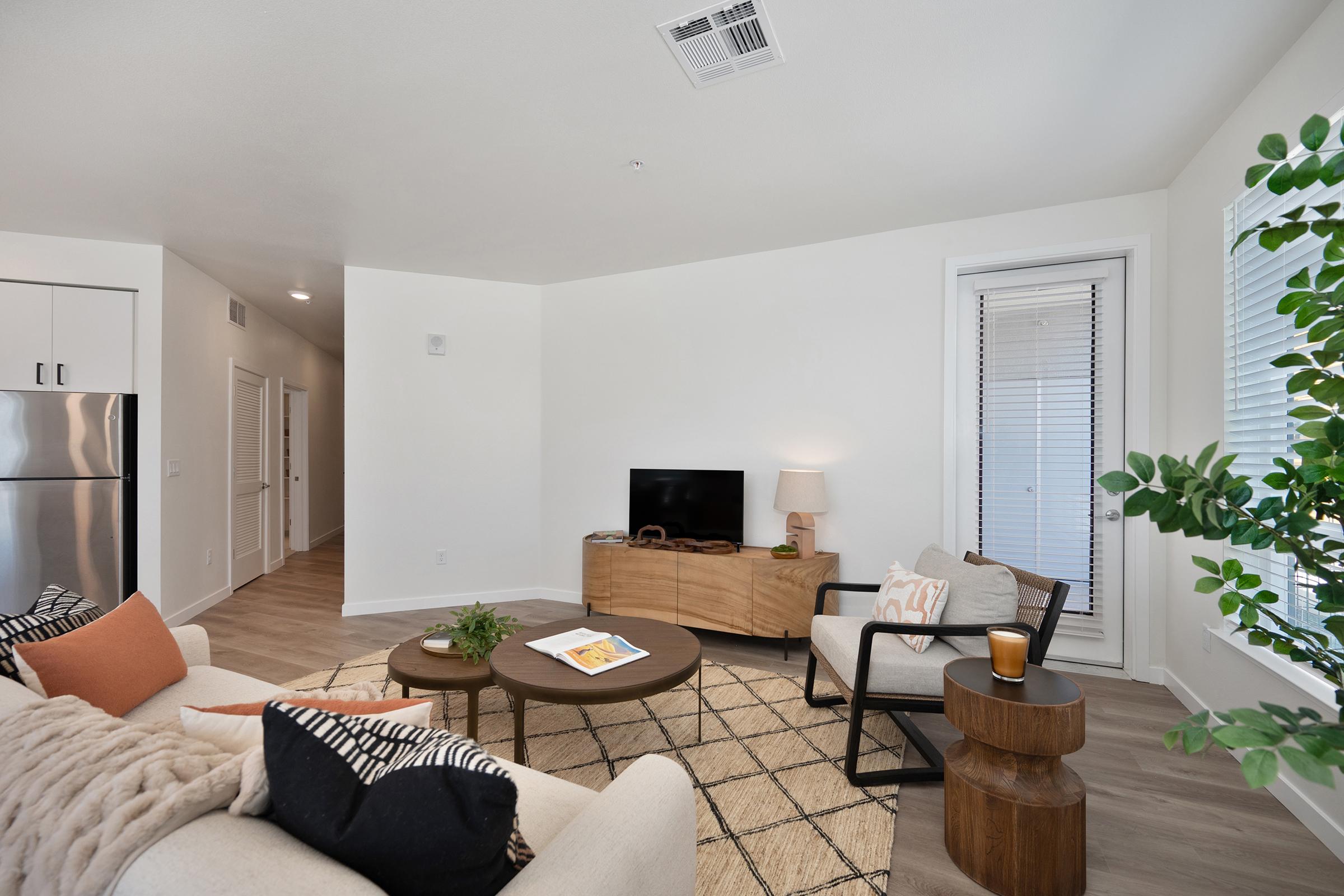 A cozy living room featuring a light beige sofa with decorative pillows, a round coffee table, and a small side table. There's a television on a wooden media console, a floor lamp beside the sofa, and a large window with white blinds. Green plants add a touch of greenery to the space, enhancing the inviting atmosphere.