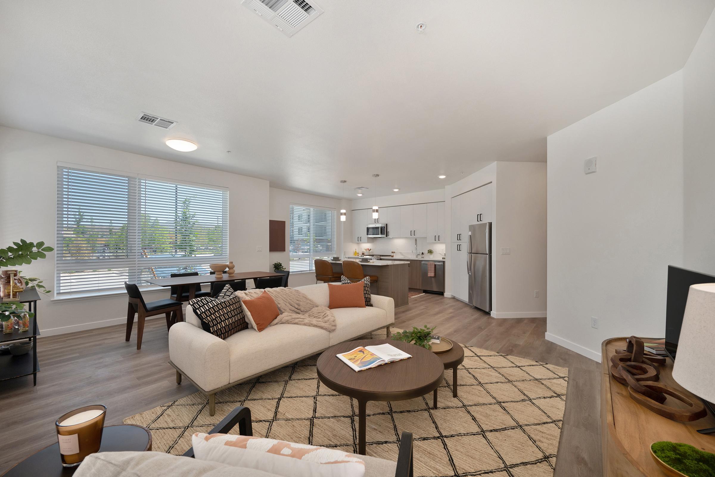 A modern, open-concept living space featuring a light-colored couch with decorative pillows, a coffee table on a patterned rug, and a dining area with dark wood chairs. Large windows provide natural light, while a contemporary kitchen with sleek appliances is visible in the background.