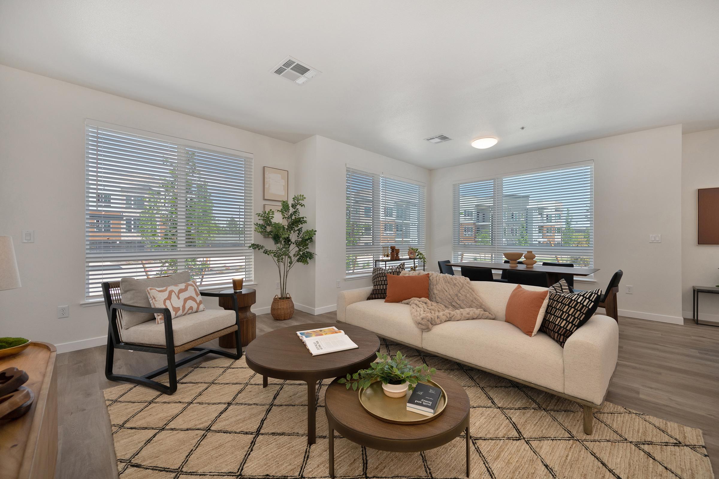 A modern living room featuring a light-colored sofa with decorative pillows, a round coffee table, and a cozy armchair. Large windows allow natural light to fill the space, complemented by indoor plants and tasteful decor. A dining area with a dark table is visible in the background, creating a warm and inviting atmosphere.