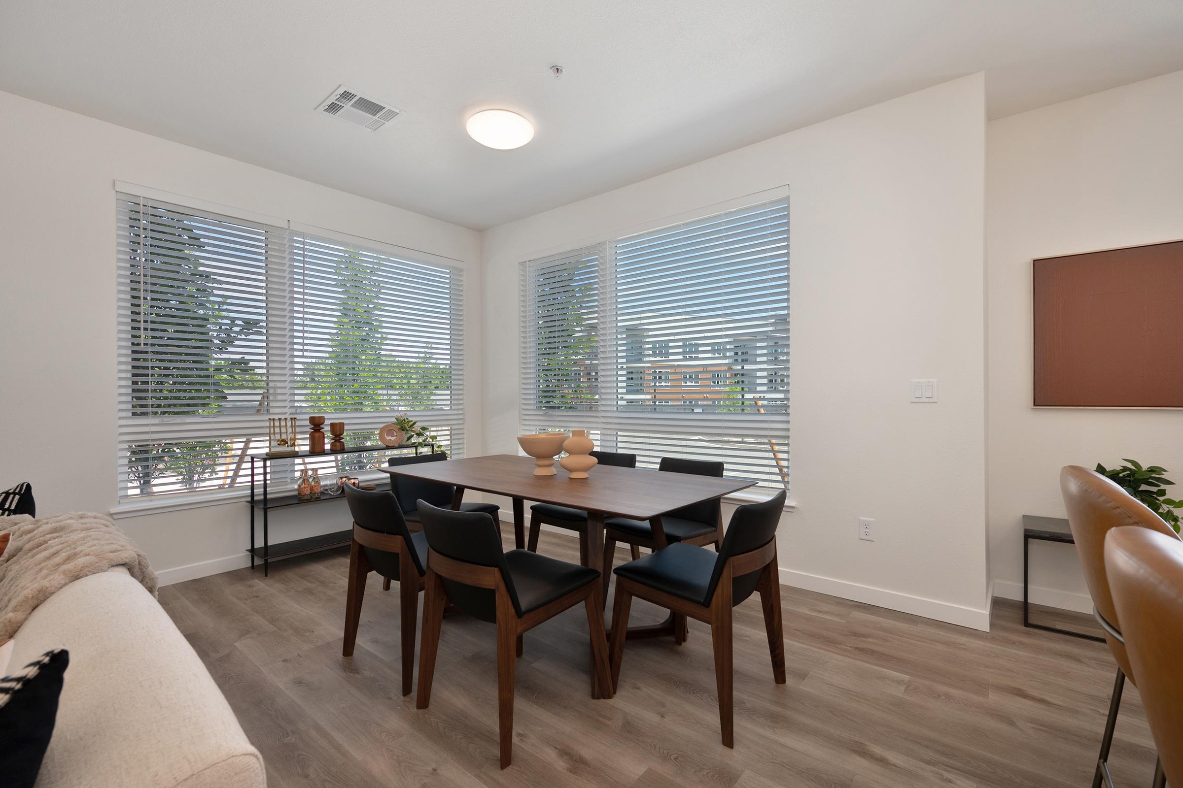 A modern dining area featuring a wooden table surrounded by black chairs, with large windows covered by blinds allowing natural light. Decorative items are placed on the table, and a couch sits nearby, with a plant in the corner and a wall art piece in a neutral tone. The space has a clean and minimalist design.
