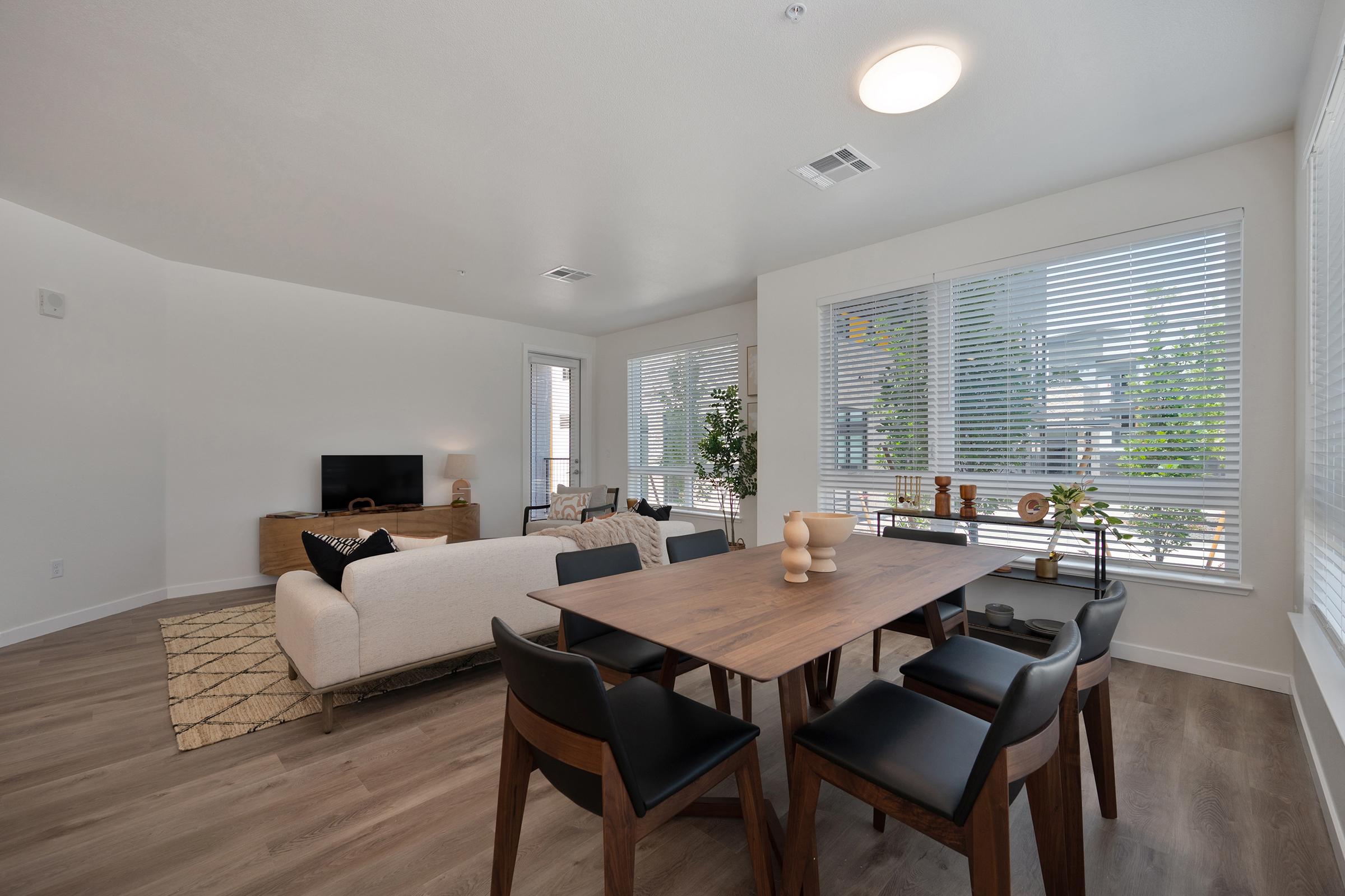 A modern living and dining room featuring a stylish sofa, a wooden dining table with black chairs, and a flat-screen TV on a side console. Large windows allow natural light, adorned with minimalist decor and greenery for a fresh ambiance. The floor is made of light wood, enhancing the contemporary feel.