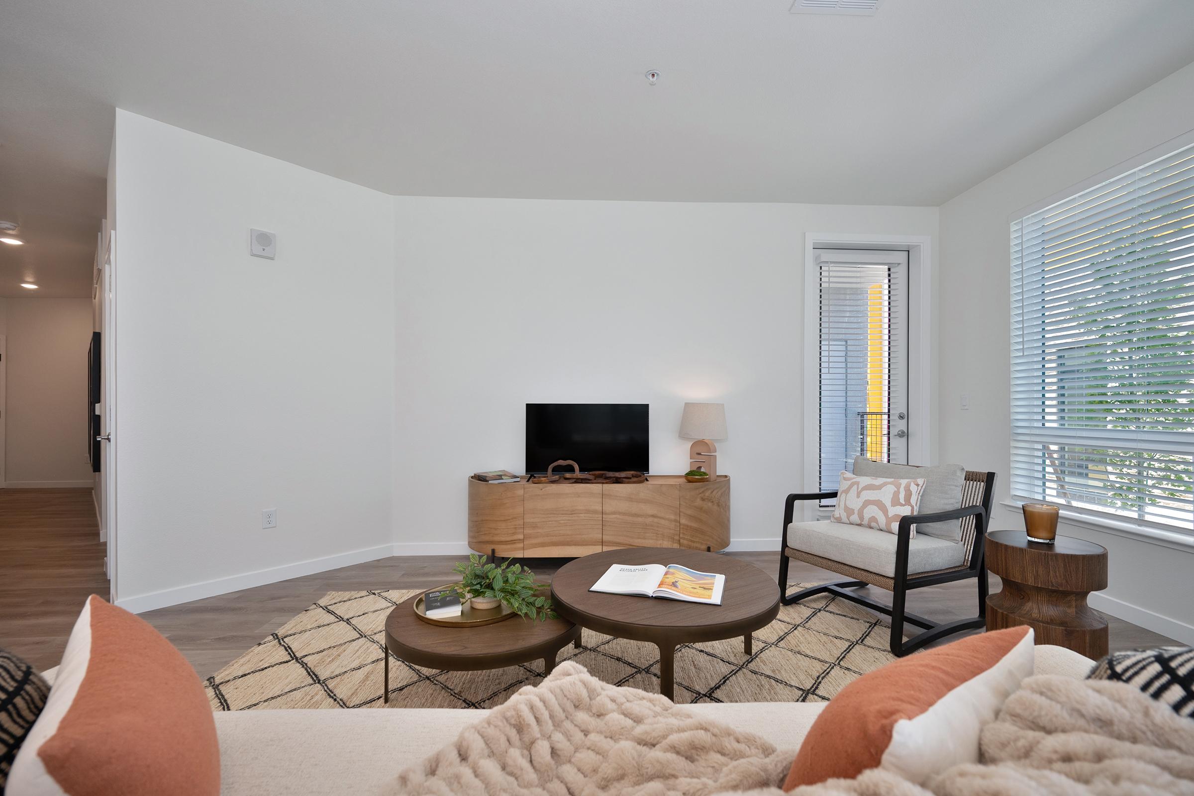 A cozy living room featuring a light-colored sofa with decorative pillows, a round coffee table, and a flat-screen TV on a wooden media console. Natural light streams in through a window with blinds, enhancing the warm ambiance of the space.