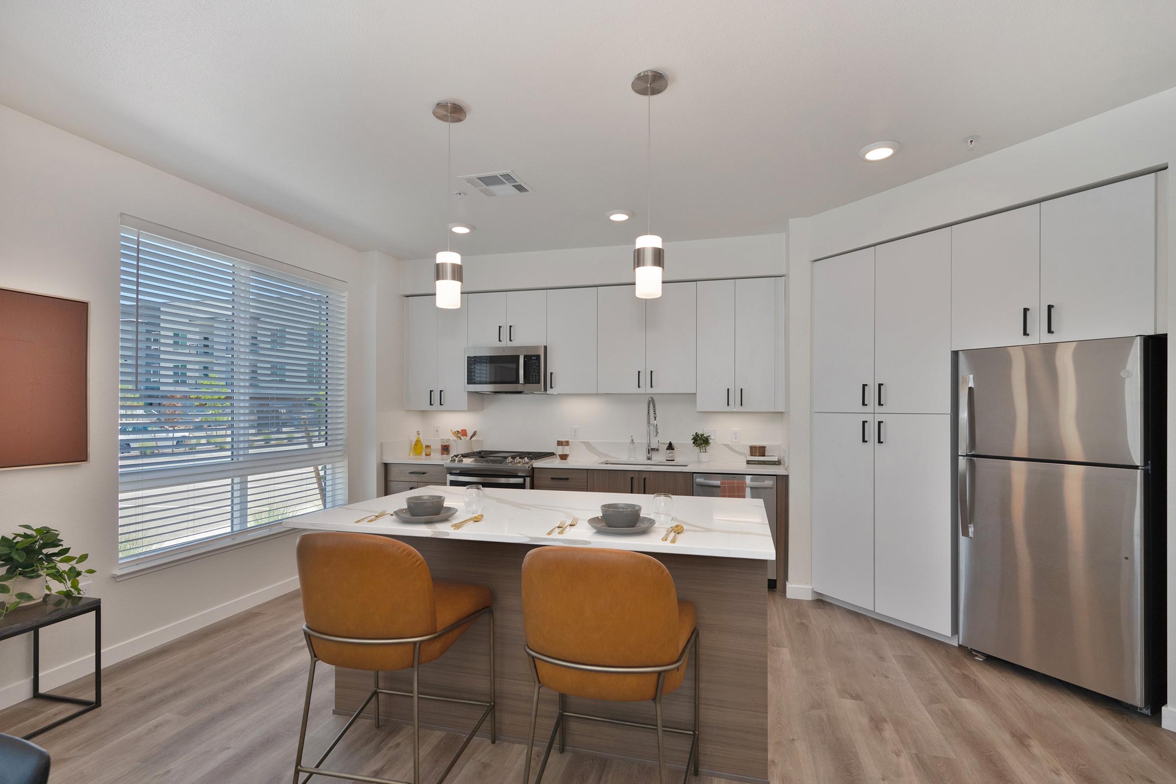 Modern kitchen featuring light cabinetry, stainless steel appliances, and a large island with seating for two. Ample natural light comes through the window, highlighting the stylish decor. The space includes gray countertops and a sleek refrigerator, creating a contemporary atmosphere.
