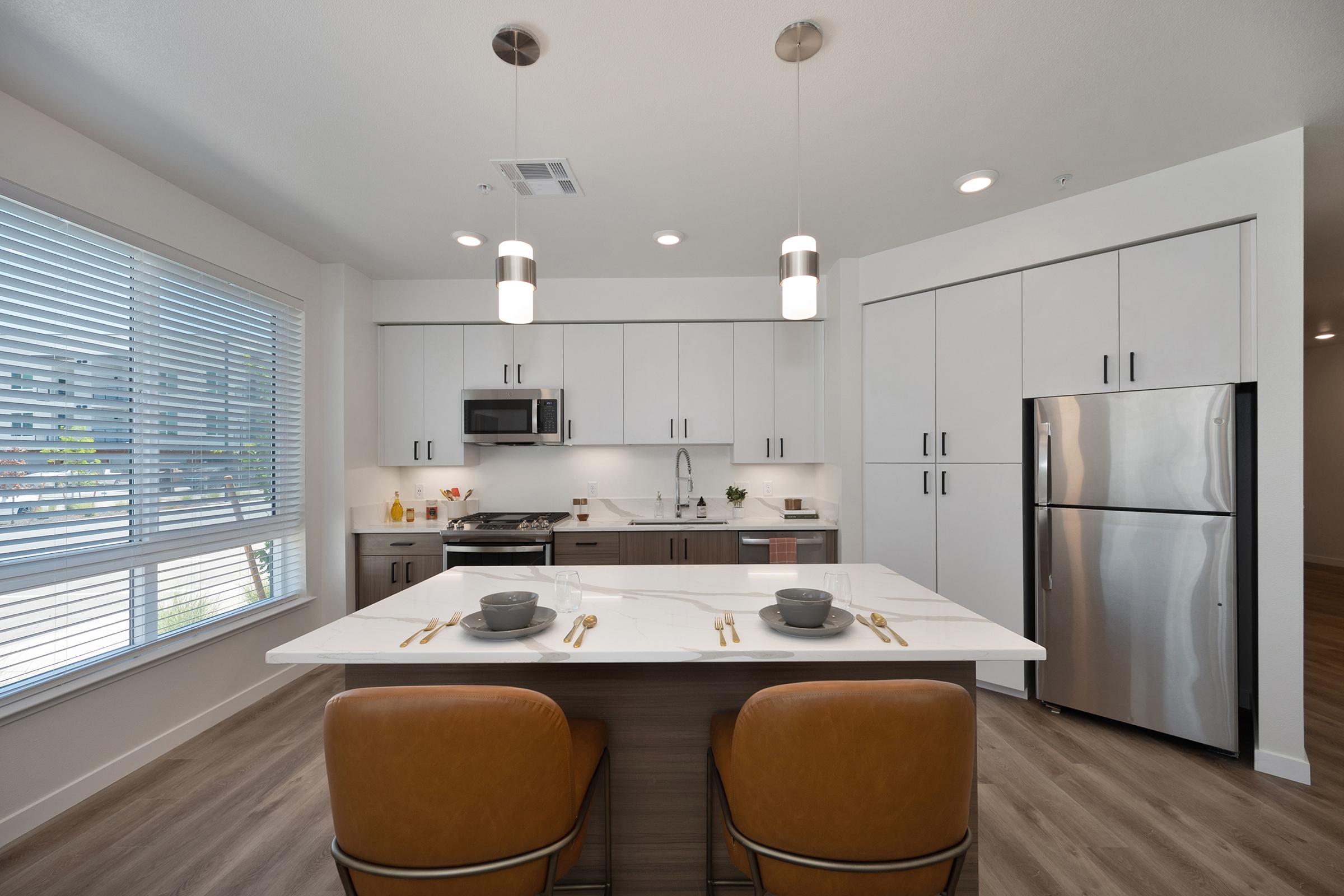 Modern kitchen featuring sleek white cabinetry, stainless steel appliances, and a large island with two brown leather chairs. Natural light streams in through large windows, illuminating the space. Table settings include grey bowls and gold utensils, creating an inviting atmosphere.