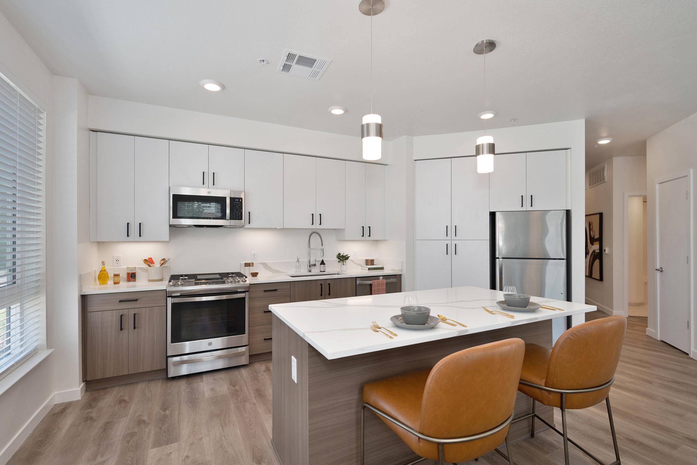 Modern kitchen featuring a central island with two brown leather bar stools, white cabinets, stainless steel appliances, and a stylish countertop. Bright lighting and large windows enhance the spacious feel, with minimalistic decor and a neutral color palette.