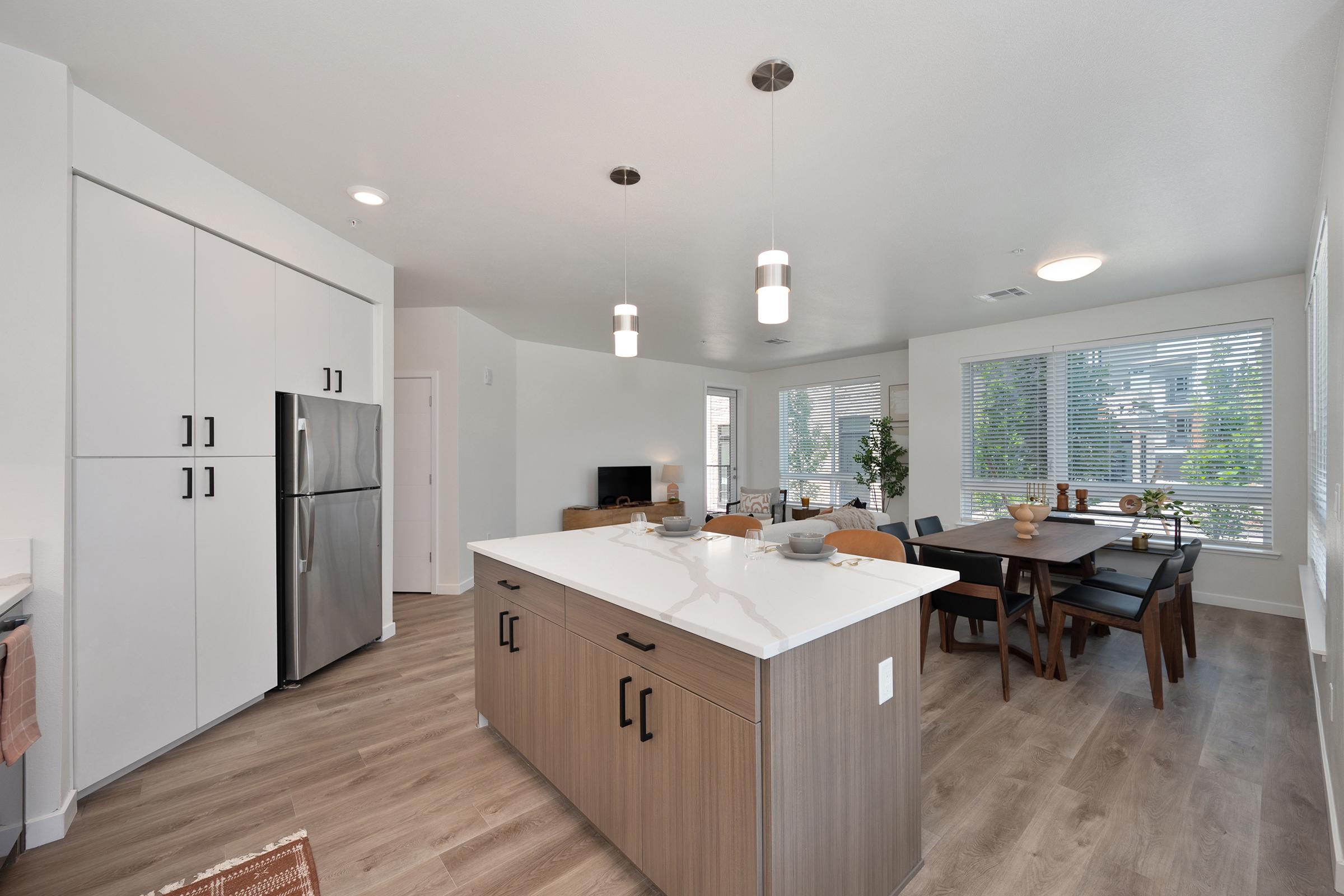 A modern kitchen and dining area featuring an island with a white countertop, dark wood cabinets, and stainless steel appliances. The dining table is set for six, with natural light streaming in through large windows. The space has a contemporary design with neutral colors and stylish lighting fixtures.