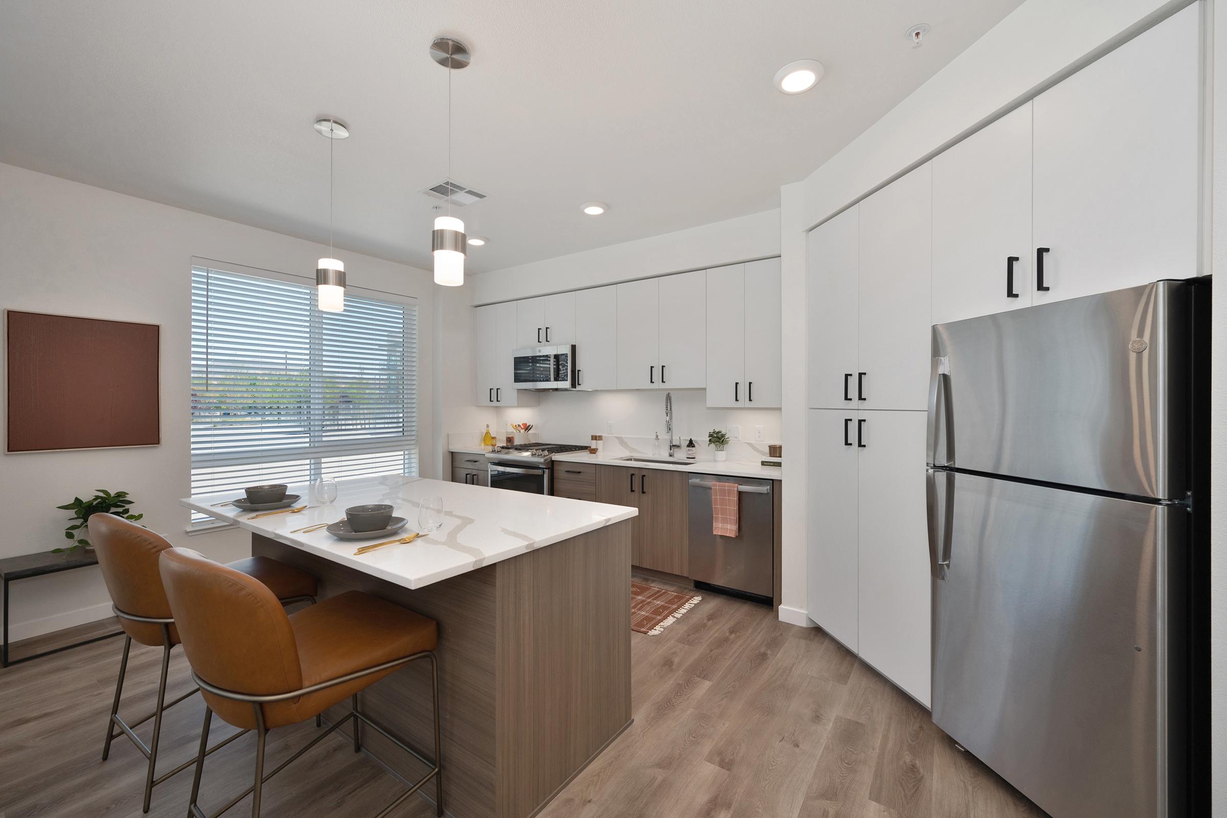 Modern kitchen with sleek white cabinets and stainless steel appliances. A central island features bar stools, perfect for casual dining. Natural light enters through a window with blinds, illuminating the space, which includes a subtle decorative piece on the wall and a patterned rug.