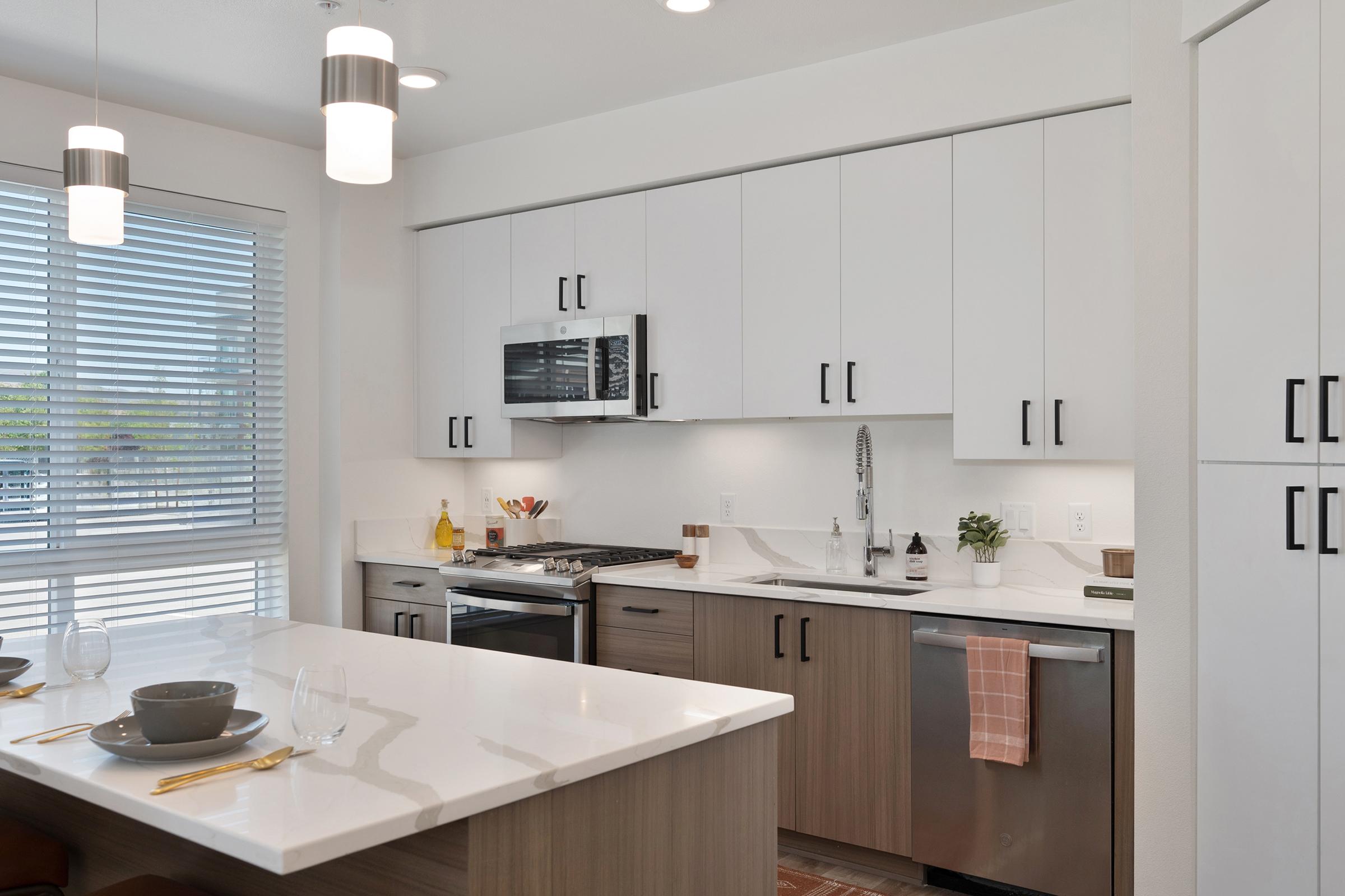 Modern kitchen featuring white cabinets with black handles, stainless steel appliances including a microwave and stove, a quartz countertop, and a spacious island. Natural light streams through a large window with blinds, creating a warm and inviting atmosphere. Accented by simple decor and dining ware set on the island.