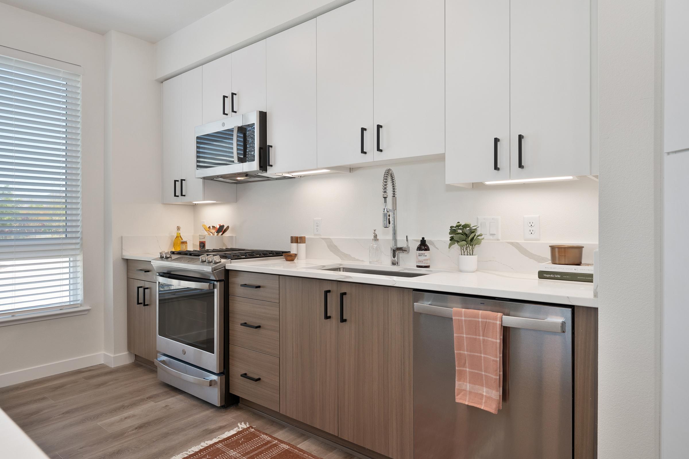 Modern kitchen featuring light wood and white cabinetry, a stainless steel gas stove, and a dishwasher. There’s a marble countertop with a small plant and kitchenware. Natural light enters through a window with blinds, and a textured rug is laid on the wooden floor.