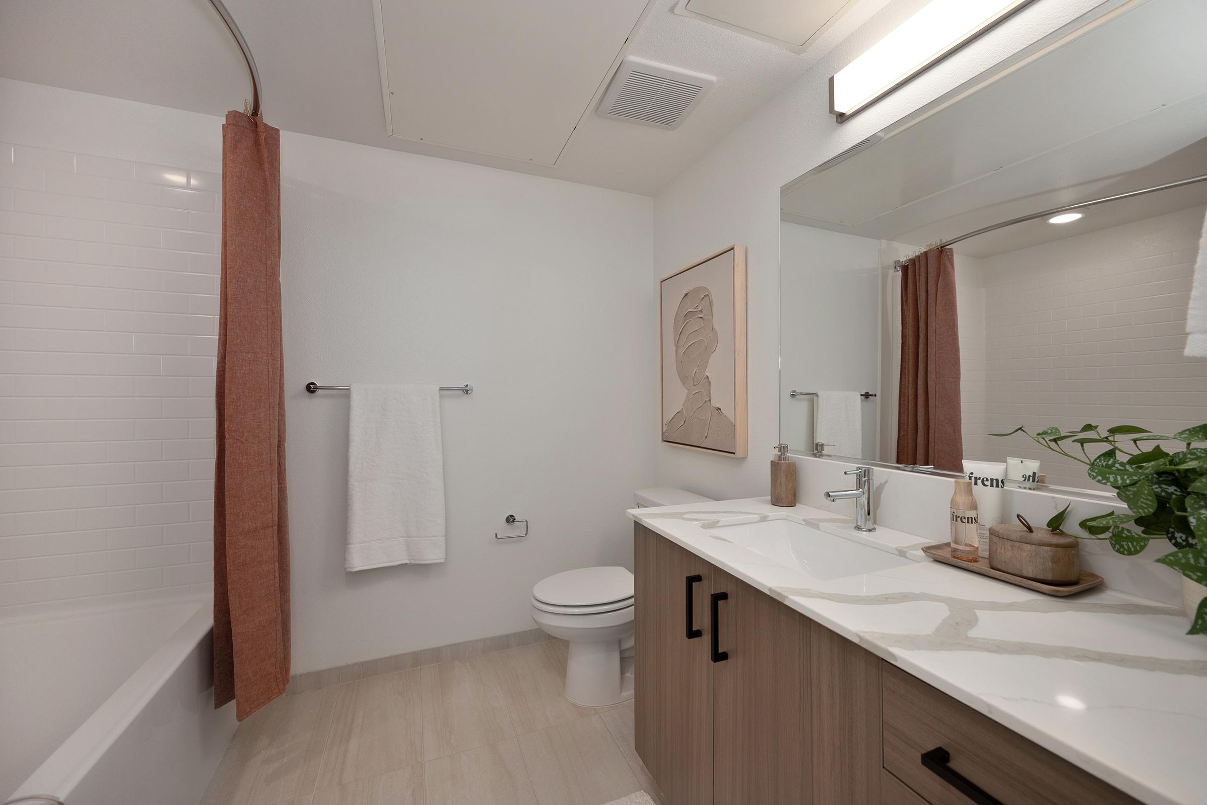 A modern, well-lit bathroom featuring a white bathtub, a large mirror, and a vanity with light wood cabinetry. A white towel hangs on a rack, and there is a decorative plant on the countertop. The walls are painted a soft color, complemented by a warm-toned shower curtain and a framed art piece.