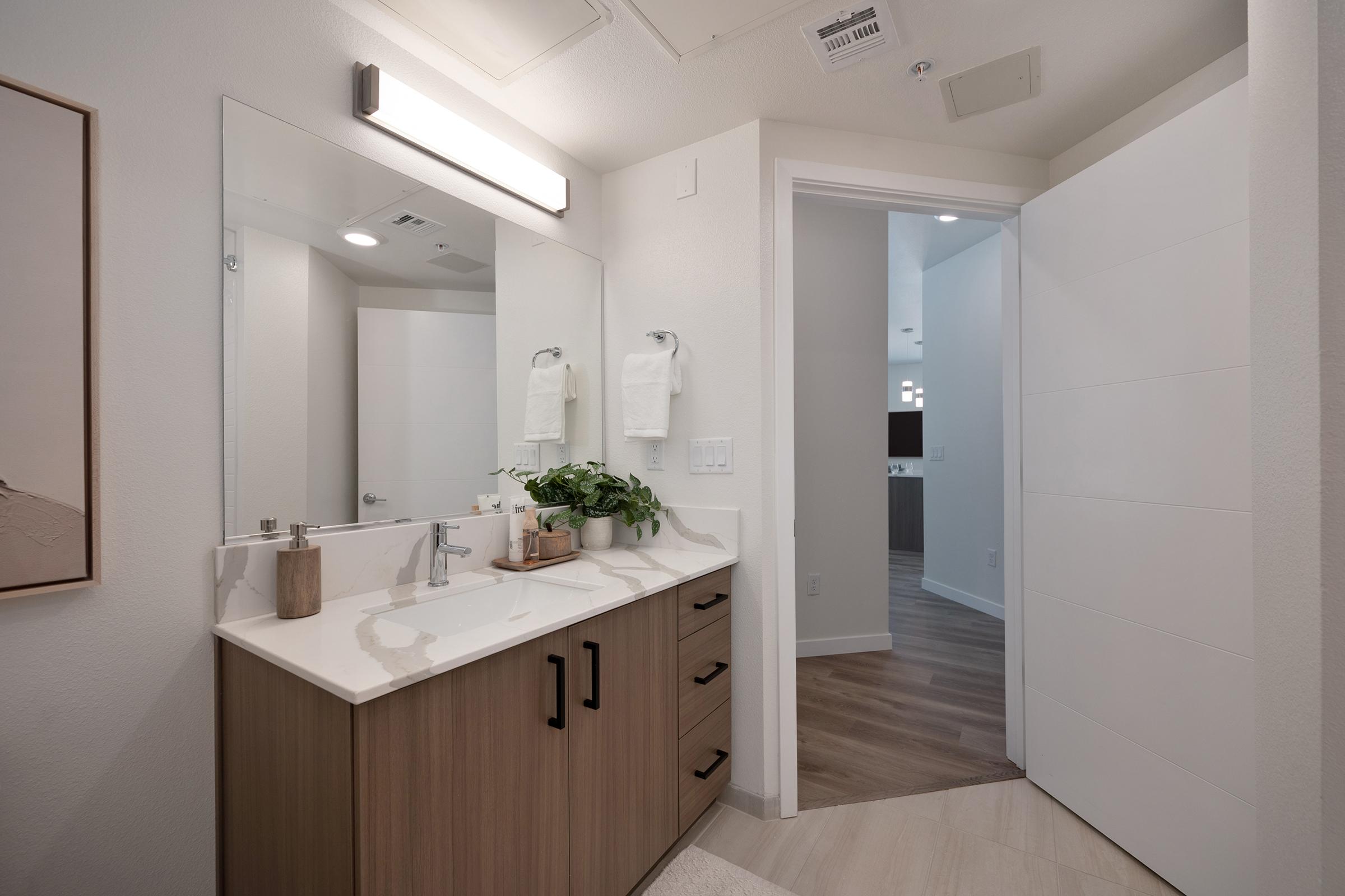 Modern bathroom interior featuring a sleek mirror above a stylish vanity with a faucet, wooden cabinetry, and decorative plants. White towels hang neatly on towel bars. A door leading to another room is visible in the background, while the floor showcases wooden planks, creating a warm and inviting atmosphere.