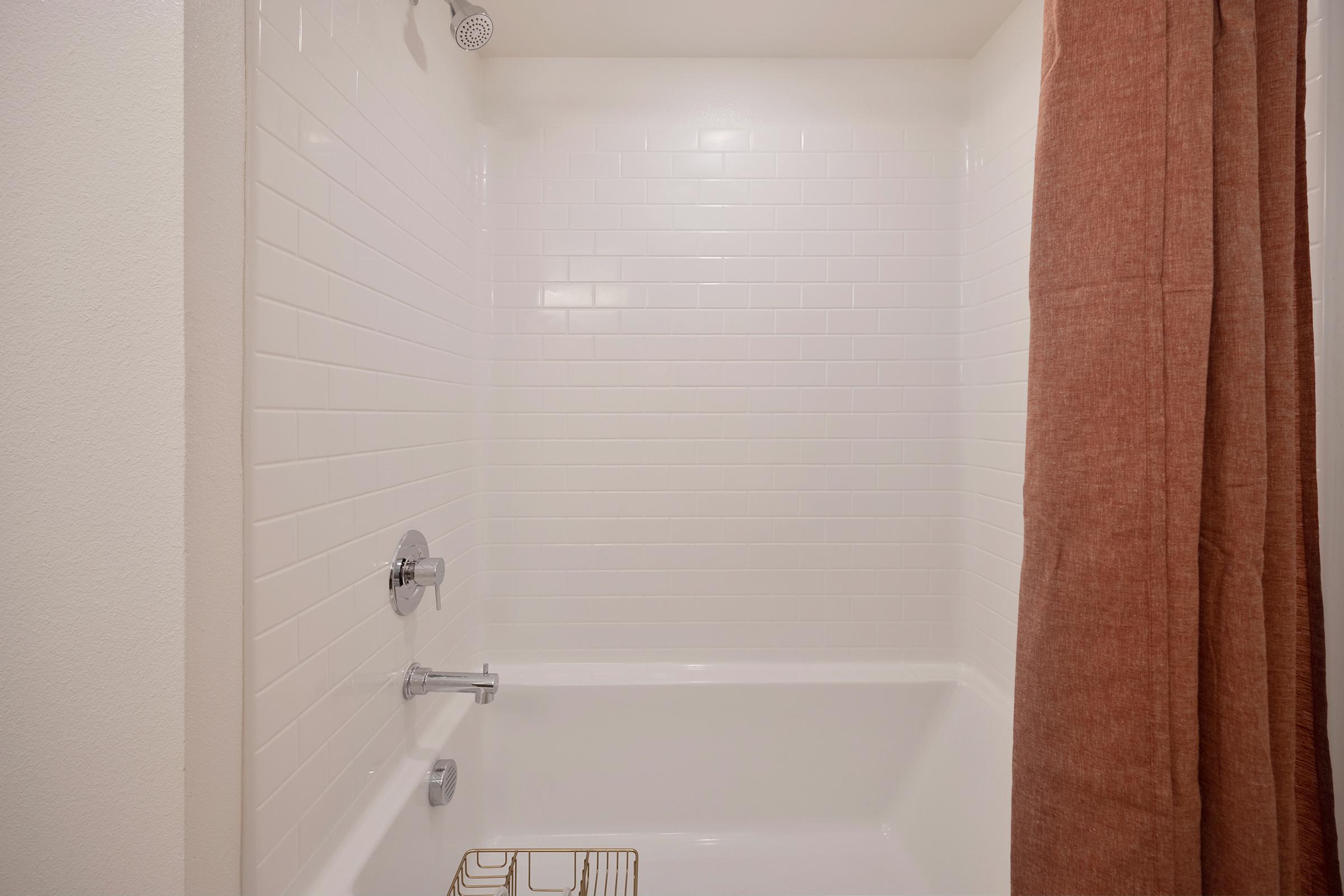 A clean, well-lit bathroom featuring a white tiled shower area with a bathtub. There is a brownish-orange shower curtain partially covering the opening, and a chrome showerhead and faucet are visible on the wall. The overall appearance is simple and modern.