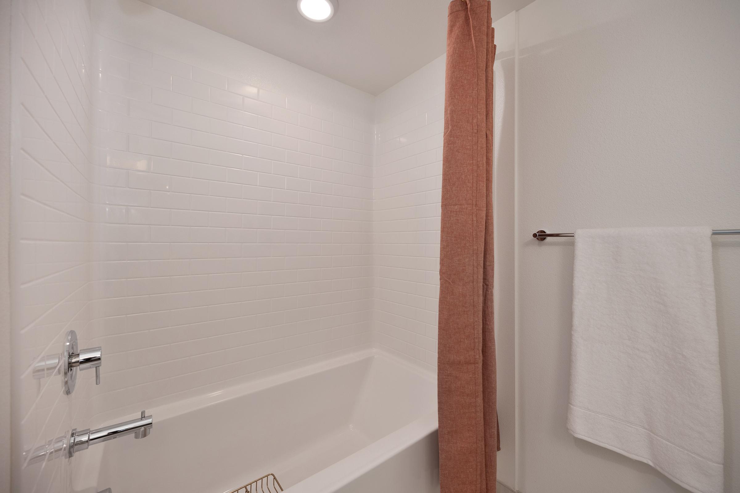 A modern bathroom featuring a white tiled shower area with a bathtub, a shower curtain in a rust color, a towel hanging on a rack, and bright overhead lighting. The clean, minimalist design creates a spacious and inviting atmosphere.