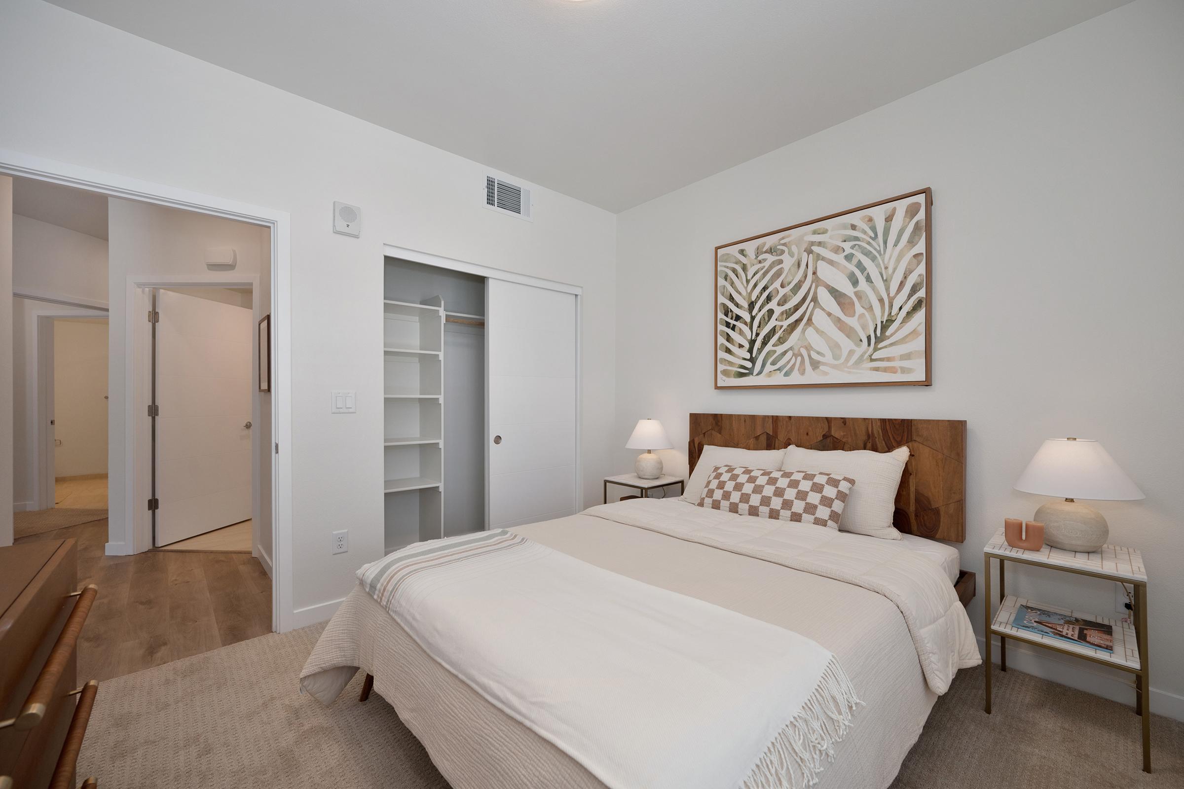 A well-decorated bedroom featuring a cozy bed with a light-colored blanket and decorative pillows. There are two bedside tables with lamps on either side, a large artwork above the bed, and a closet with sliding doors. The room has white walls and a minimalist design, creating a serene atmosphere.