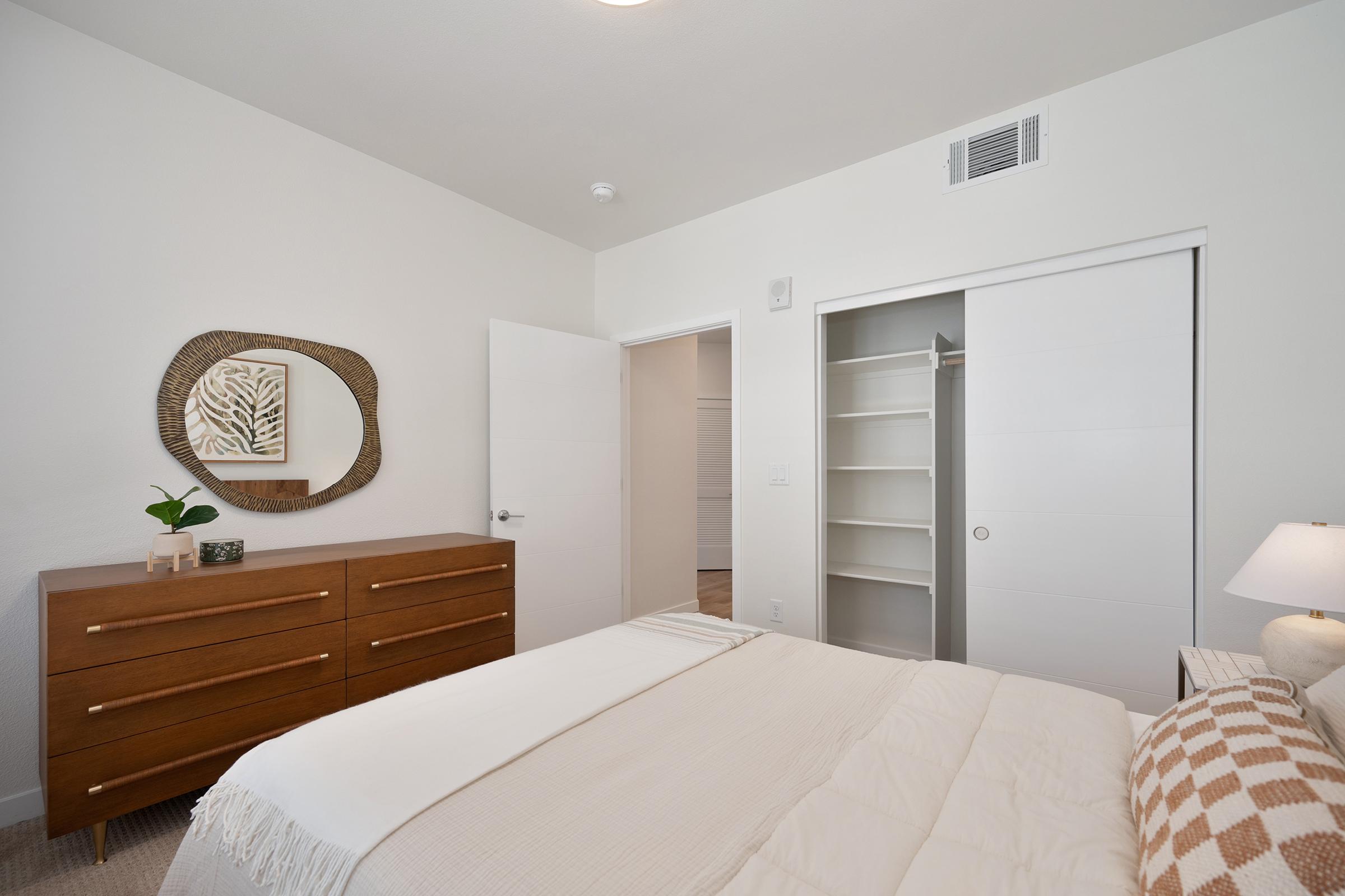 A cozy bedroom featuring a neatly made bed with a light-colored blanket, a stylish wooden dresser, and a decorative round mirror on the wall. A white closet door opens to additional storage space, while a lamp adds a warm touch. The walls are painted a soft white, creating a bright and airy atmosphere.