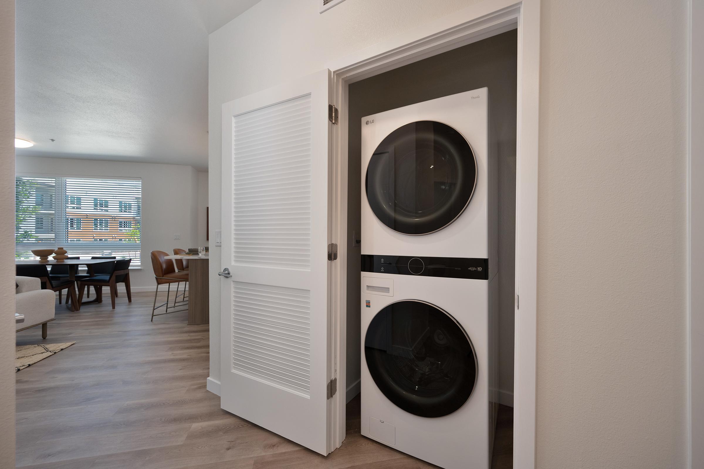 A compact laundry area featuring a stacked washer and dryer, positioned within an open doorway. The background shows a cozy living space with a dining table and chairs visible through a window, suggesting a well-designed modern apartment layout.