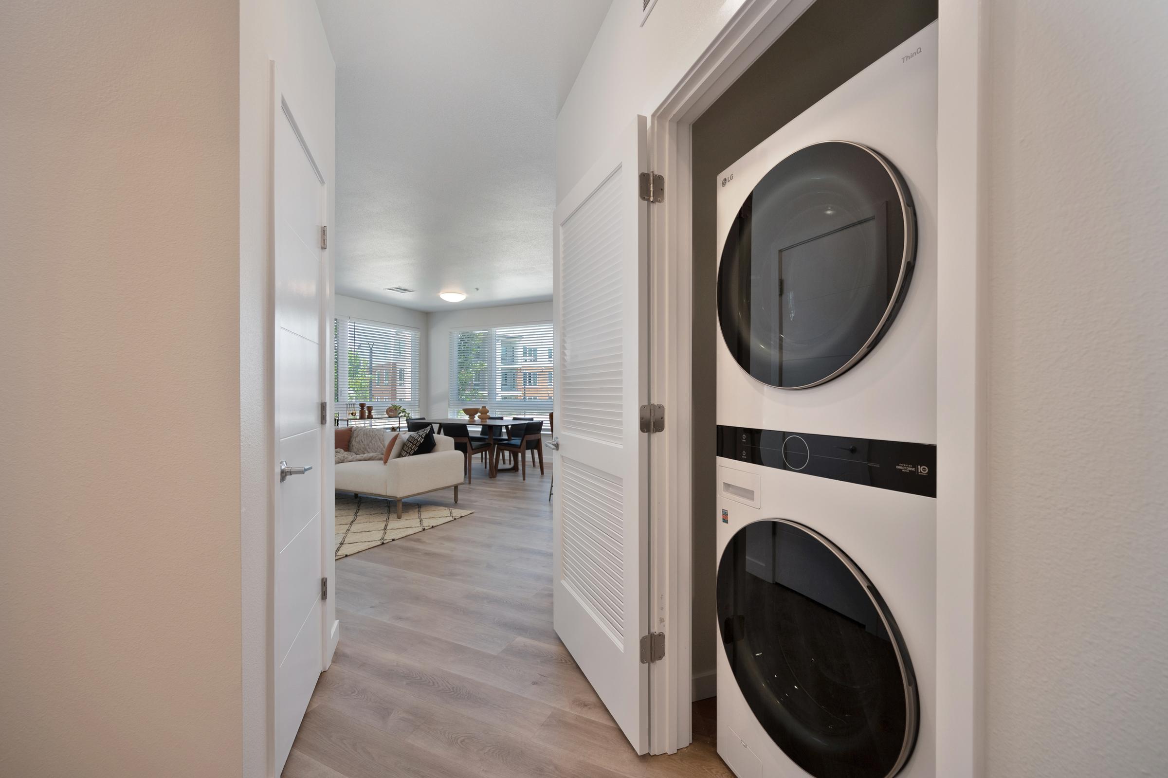 A view through an open door showcasing a modern laundry area with a stacked washer and dryer on the left. In the background, a stylish living and dining space is visible, featuring large windows, a comfortable sofa, and a dining table, all in a bright and inviting atmosphere.
