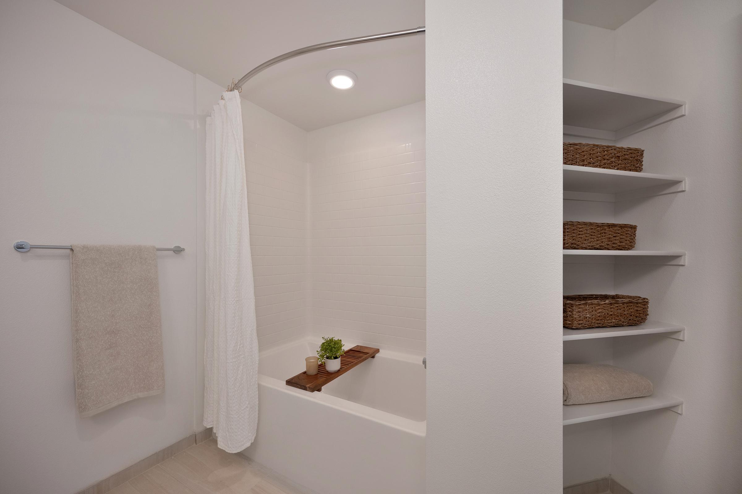 Brightly lit bathroom featuring a white bathtub with a shower curtain, a wooden shelf across the tub for decor, and neatly organized open shelves displaying woven baskets and folded towels. The walls are painted white, creating a clean and modern aesthetic.