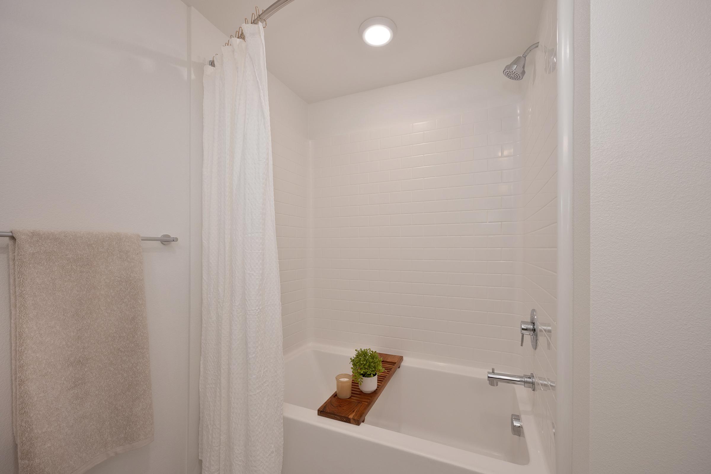 A clean, modern bathroom featuring a white tiled shower with a glass door, a white shower curtain, and a wooden bath tray holding a small potted plant and a candle. A beige towel hangs on a rack nearby, providing a cozy touch to the minimalist design. The space is well-lit with a ceiling light.