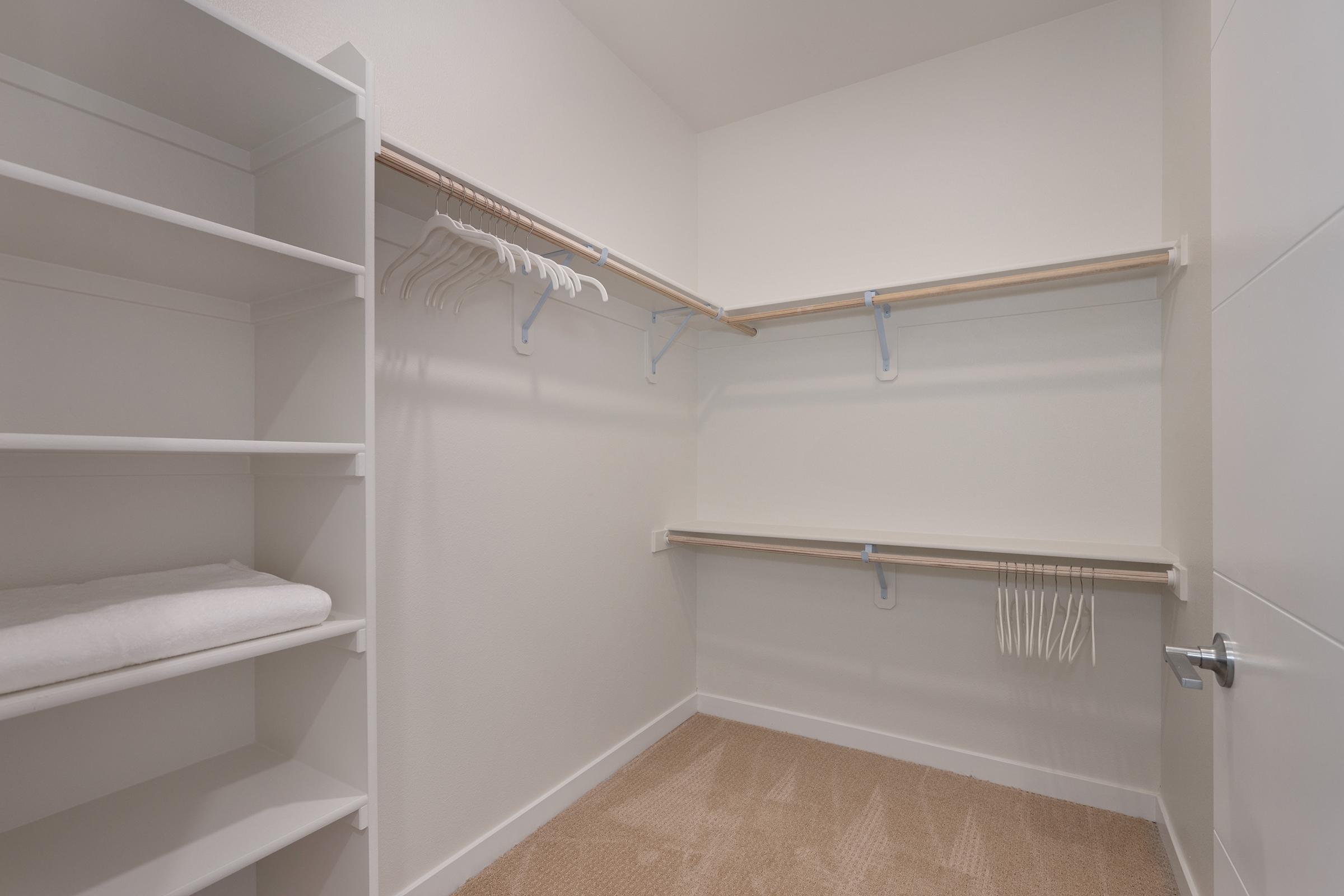 A spacious, empty walk-in closet with white walls, two wooden shelves mounted on the walls, and a section with hanging rods. There is a stack of neatly folded towels on one shelf and a beige carpeted floor, creating a clean and organized look.