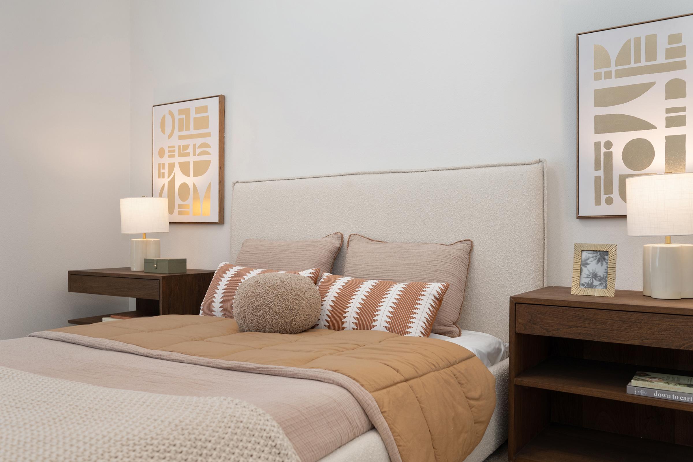Cozy bedroom with a neutral color palette featuring a beige upholstered headboard, soft throw pillows in earthy tones, and a throw blanket. Two bedside tables with stylish lamps flank the bed, and decorative wall art adds a modern touch to the space.