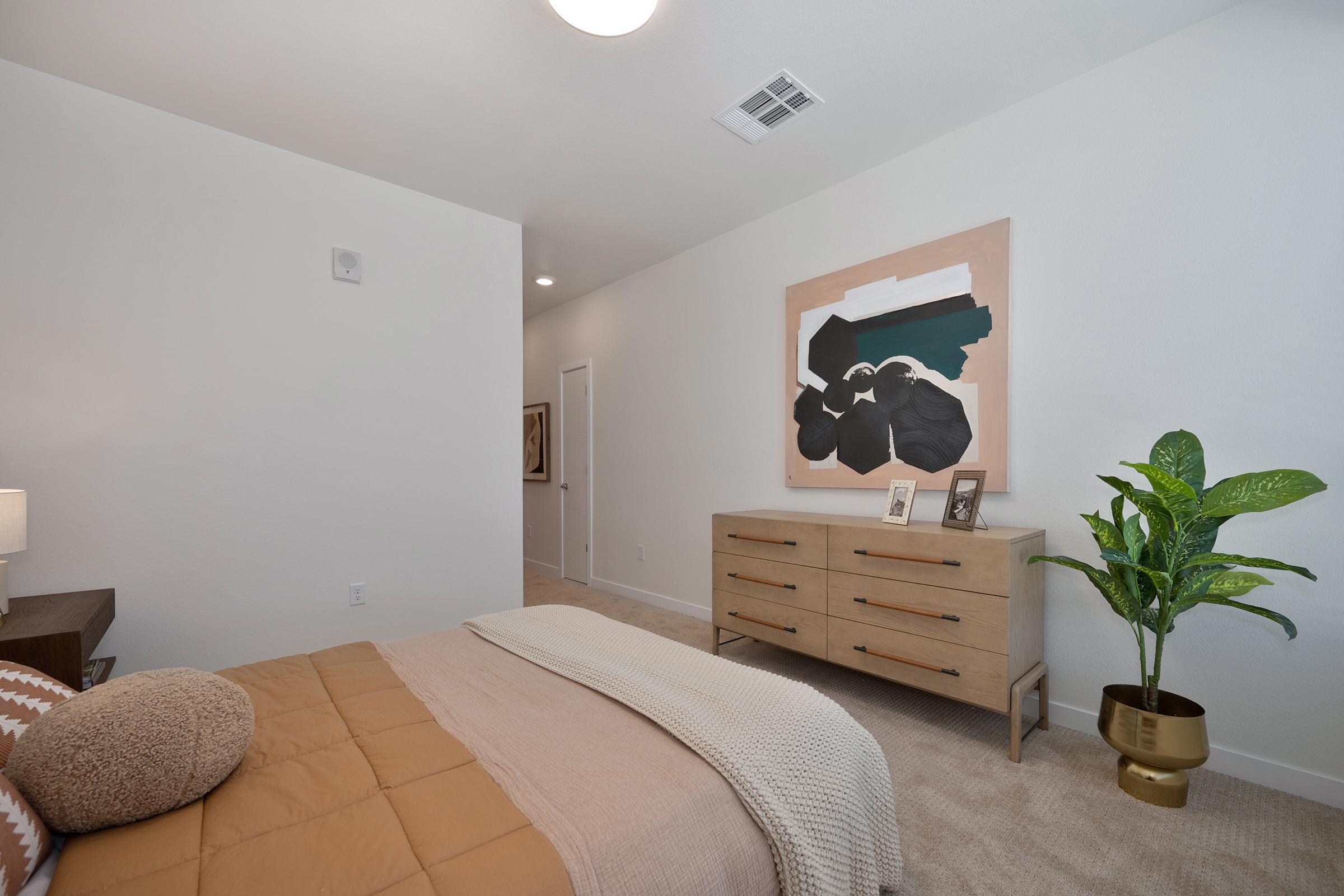 A modern bedroom featuring a neatly made bed with a beige quilt and decorative pillows. There's a wooden dresser with framed photos and a potted plant. A large abstract painting hangs above the dresser, and the room has neutral-colored walls and carpet, creating a cozy, inviting atmosphere.