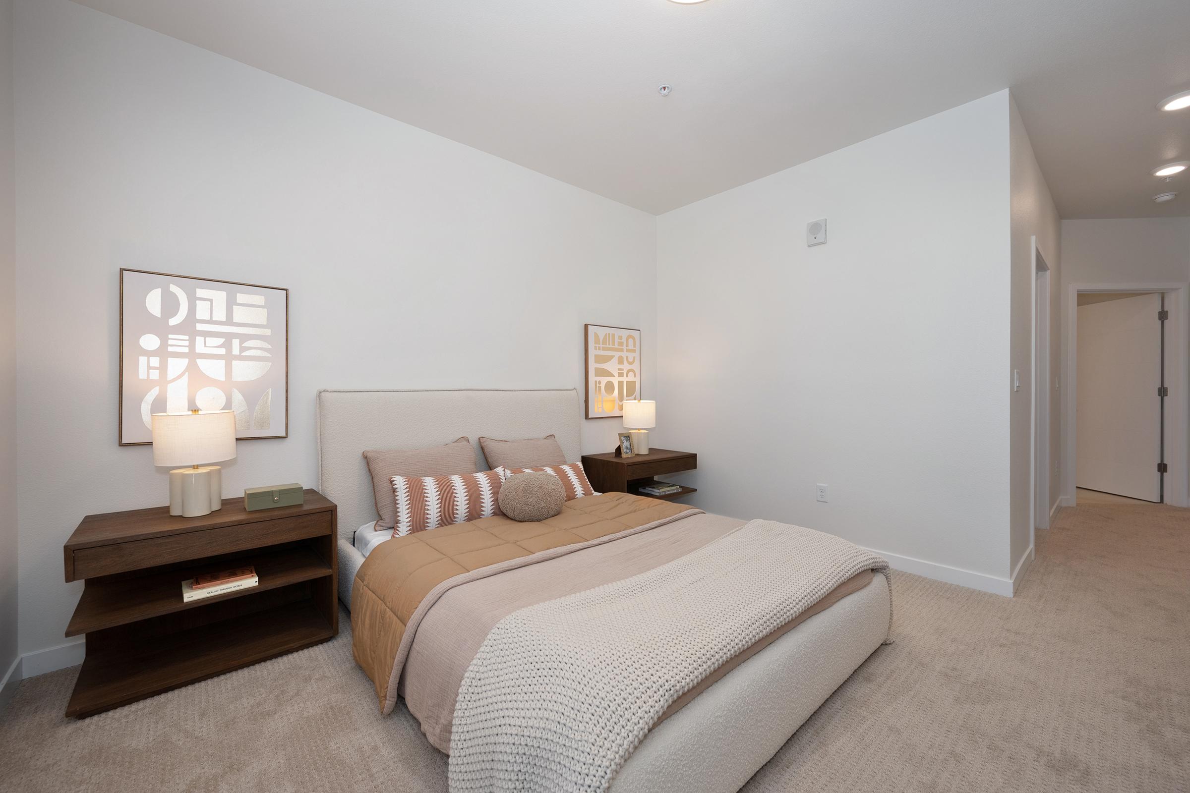 A modern bedroom featuring a beige bedspread and decorative pillows on a bed with a light-colored headboard. On either side of the bed, there are wooden nightstands with lamps. The walls are painted white, and there are framed artworks above the nightstands. The room has neutral carpeting and a minimalist design.