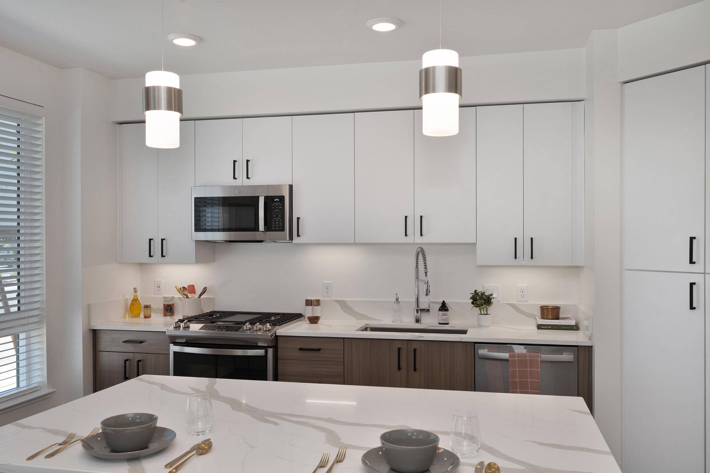 Modern kitchen with sleek white cabinetry and stainless steel appliances, including a microwave and oven. A large island in the center features a white countertop with two gray plates and glasses. The space is well-lit with pendant lights hanging above, and there are decorative items on the counter. Large windows provide natural light.