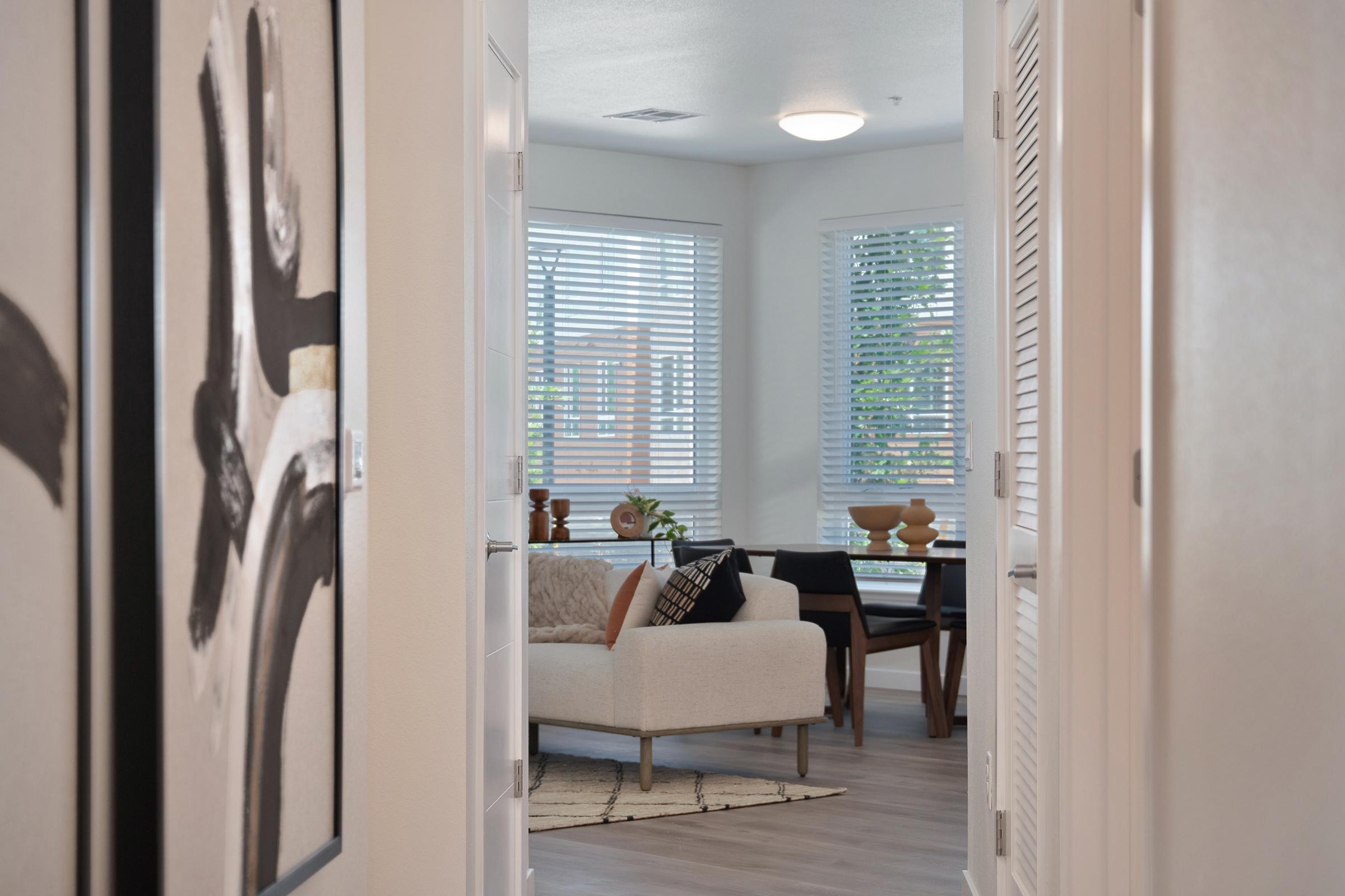 A hallway leading into a bright living area, featuring a light-colored sofa with decorative pillows, a dining table with wooden decor, and large windows with blinds letting in natural light. The walls are adorned with abstract art, and a rug is placed on the floor, enhancing the cozy atmosphere.