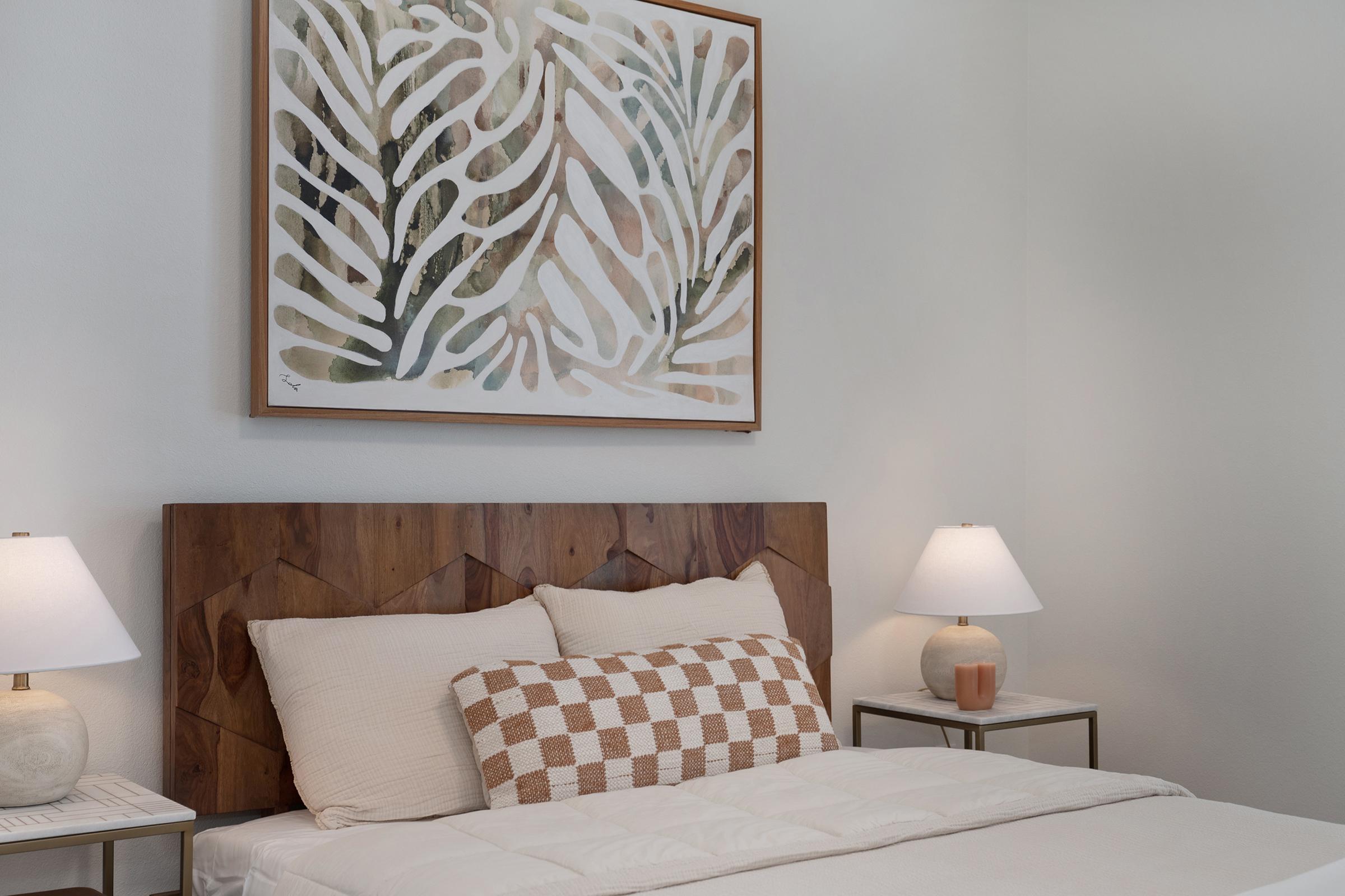 A cozy bedroom featuring a wooden headboard with a geometric design, a light-colored bed with a patterned throw pillow, and two bedside lamps. Above the bed, there is a large abstract wall art piece in earthy tones depicting leaves. The walls are painted a soft white, creating a serene atmosphere.