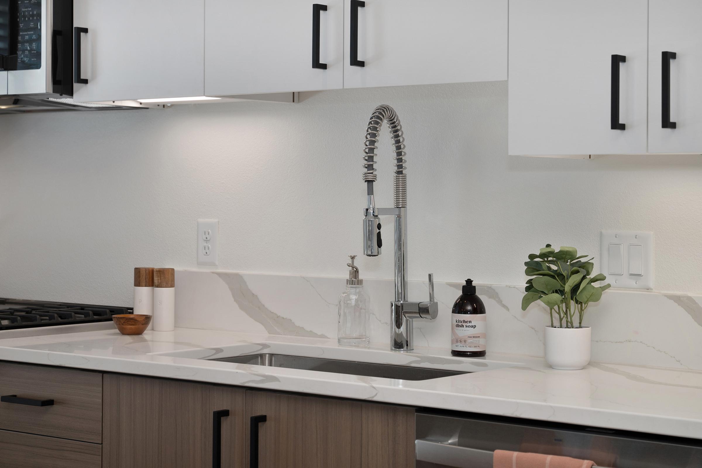 Modern kitchen with a sleek white and wood design. Features a stainless steel sink with a pull-down faucet, white cabinets with black handles, and a marble countertop. Decorative items include a small potted plant, a clear glass container, and a dark bottle on the counter.
