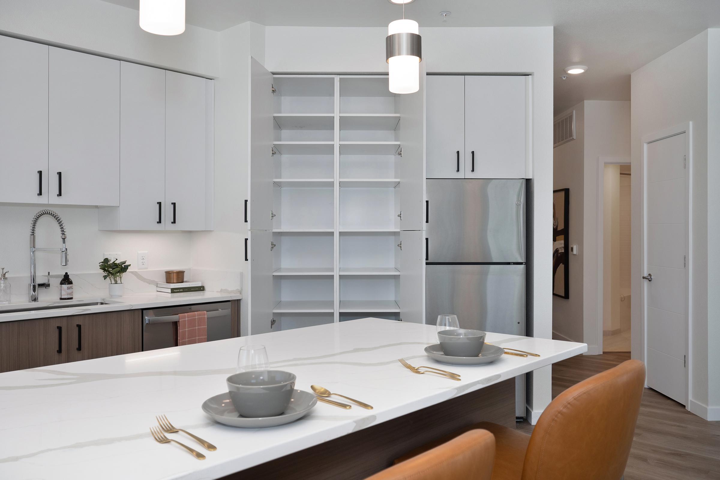 A modern kitchen featuring a large island with two gray bowls and gold utensils. The cabinetry is sleek and white, with a stainless steel refrigerator and an open shelf unit. Soft lighting from pendant fixtures enhances the contemporary design, while warm wood accents add a cozy touch.