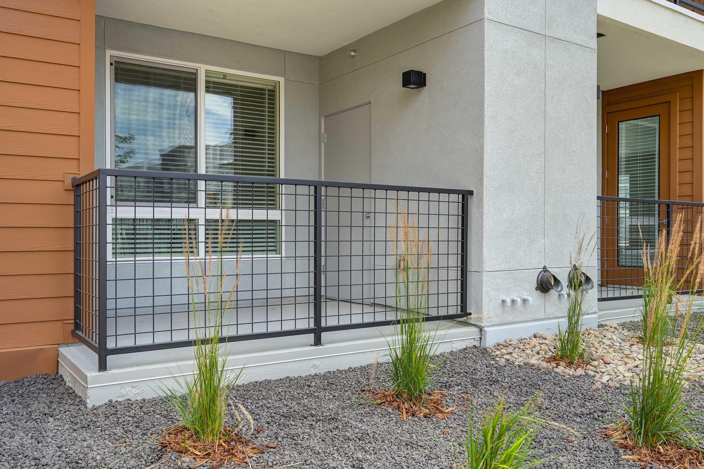 A modern entryway featuring a small covered porch with black railing, situated between two residential units. The porch has a concrete slab, and the surrounding area includes gravel landscaping with ornamental grasses and small rocks, enhancing the contemporary design.