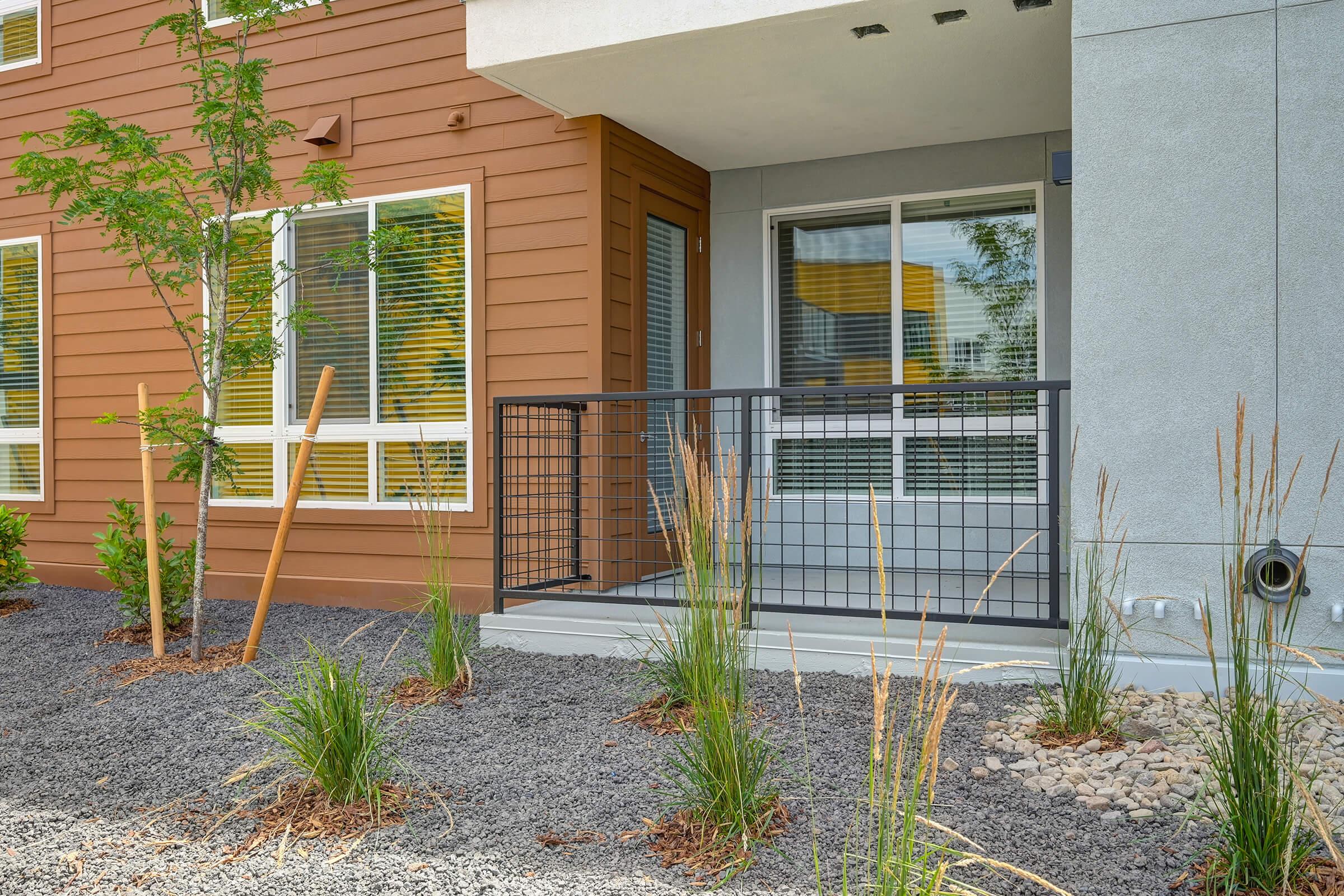 A modern building entrance featuring a metal railing, surrounded by landscaped areas with rocks and grasses. The exterior includes a mix of brown and gray materials, large windows, and minimalistic design elements. Young trees are also present to enhance the aesthetic appeal.