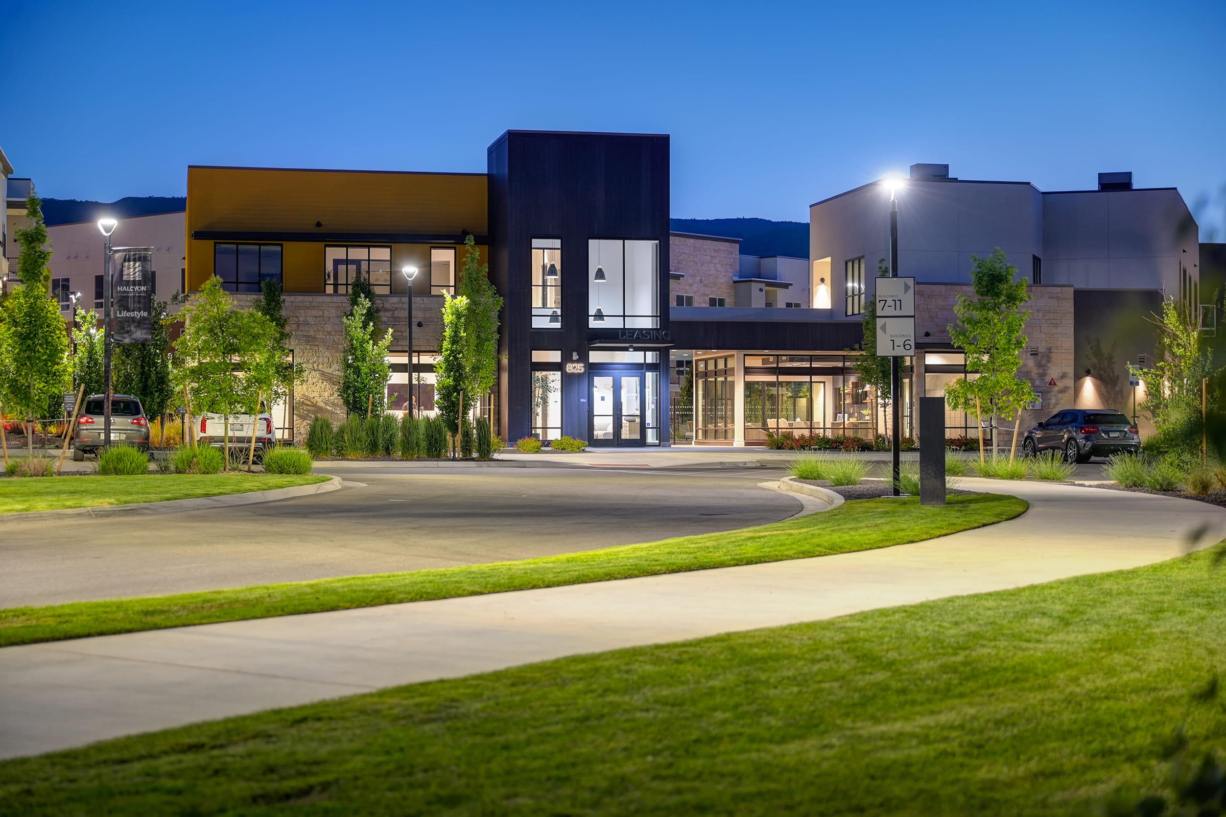 A modern building with large windows and a stylish facade, situated in a well-lit area at twilight. Surrounding the structure are trees and neatly maintained grass, while a curved pathway leads to the entrance. The scene has a clean and contemporary urban vibe.