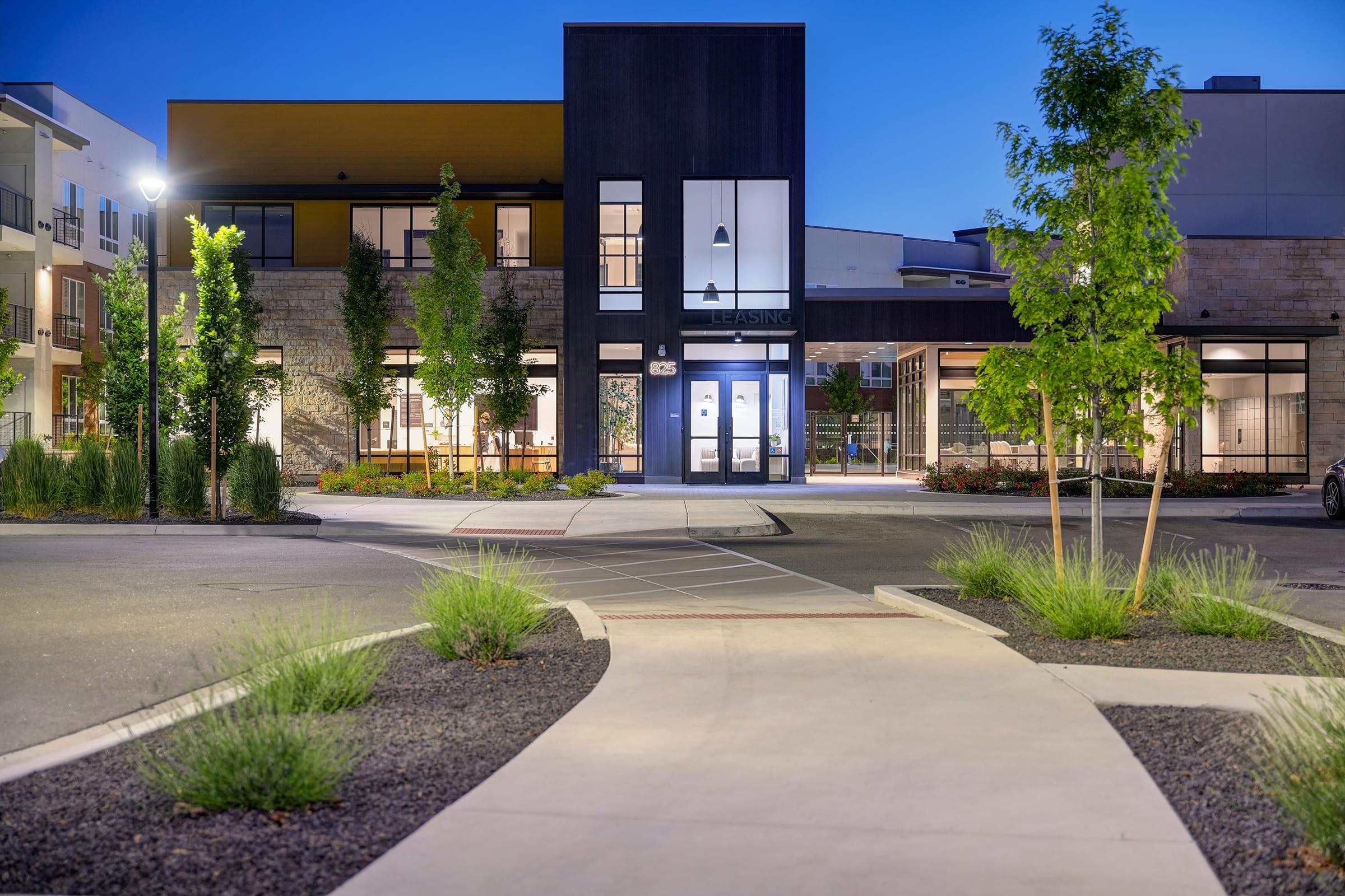 Modern building with large windows and a dark exterior, surrounded by landscaped greenery and smooth pathways. The scene is set at dusk, displaying a serene atmosphere with subtle lighting, highlighting the architectural design and surrounding plants.