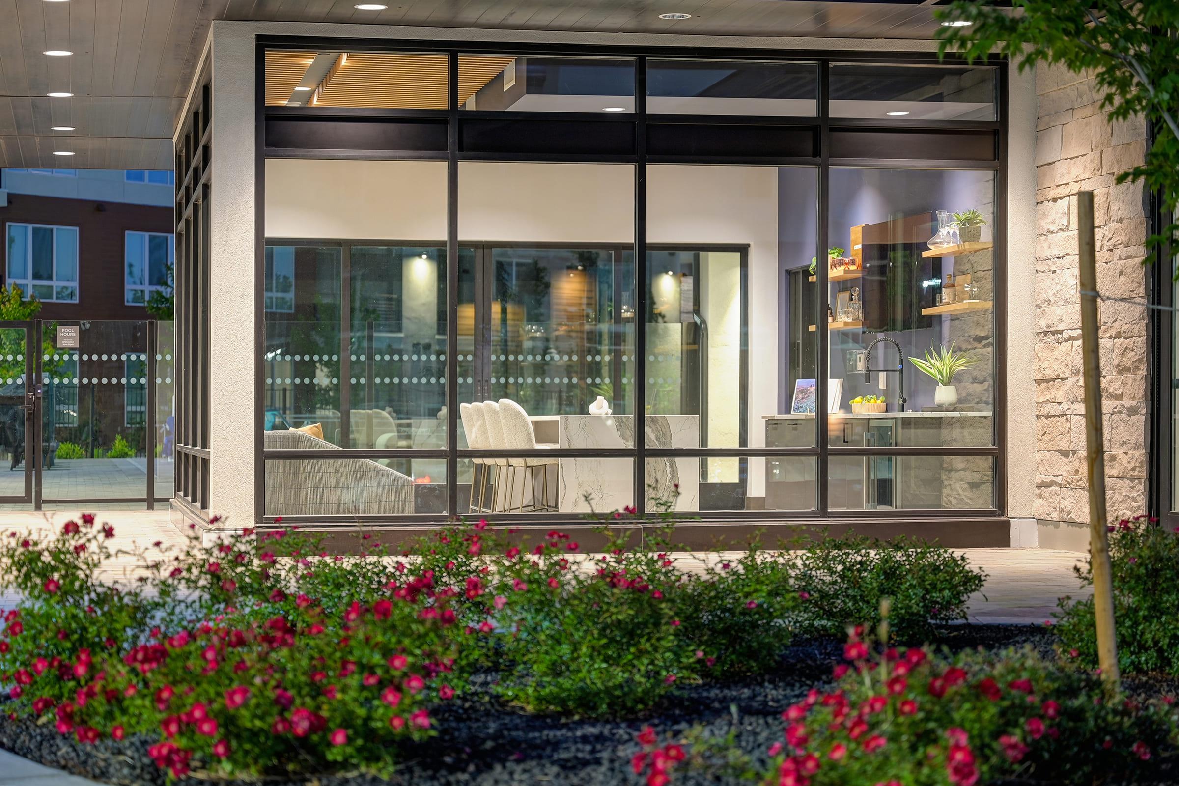 A modern building exterior featuring large glass windows that showcase a well-lit interior with stylish furniture, including white chairs and a desk. Surrounding the building are vibrant flower beds filled with pink roses, adding a touch of nature to the contemporary architecture.