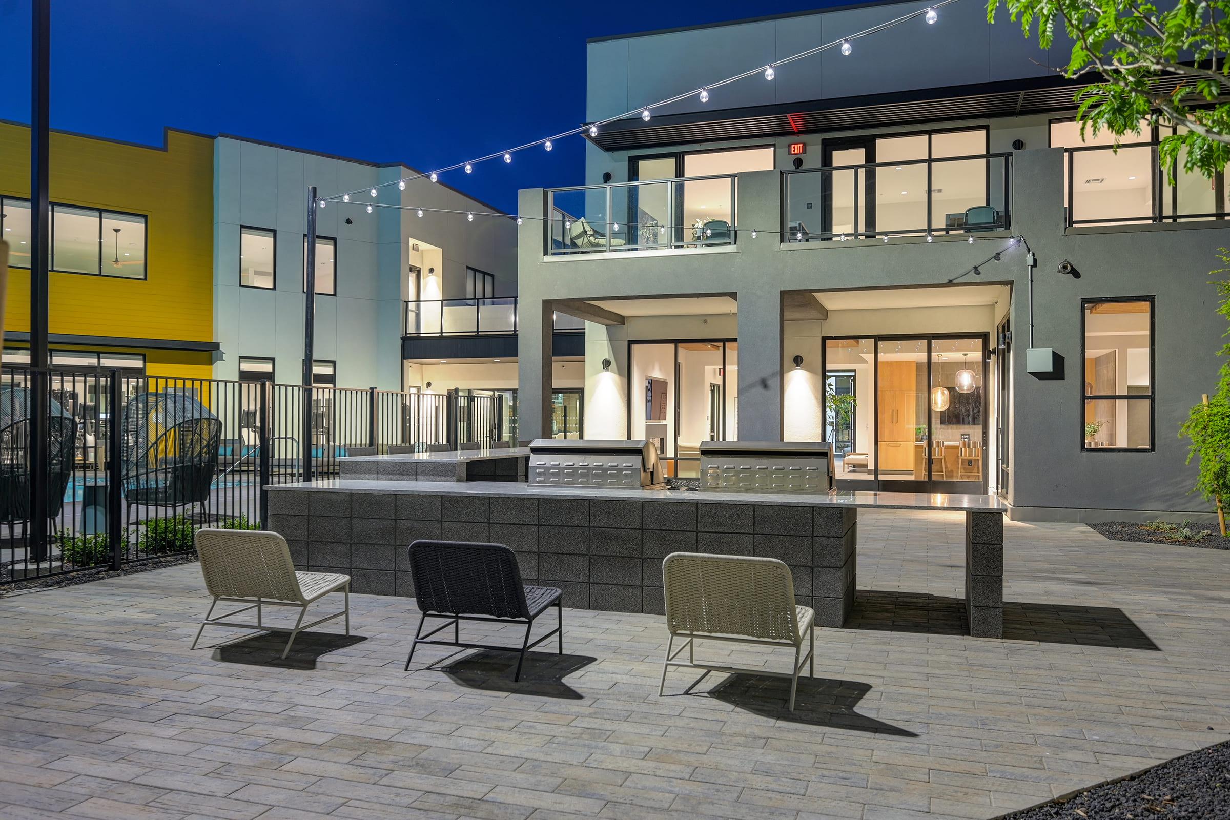 A modern outdoor space at night featuring three chairs in front of a stylish building with large windows. The area has a stone path lined with greenery and illuminated by string lights, creating a cozy atmosphere. The building's architecture is contemporary, with a balcony and an inviting entrance.